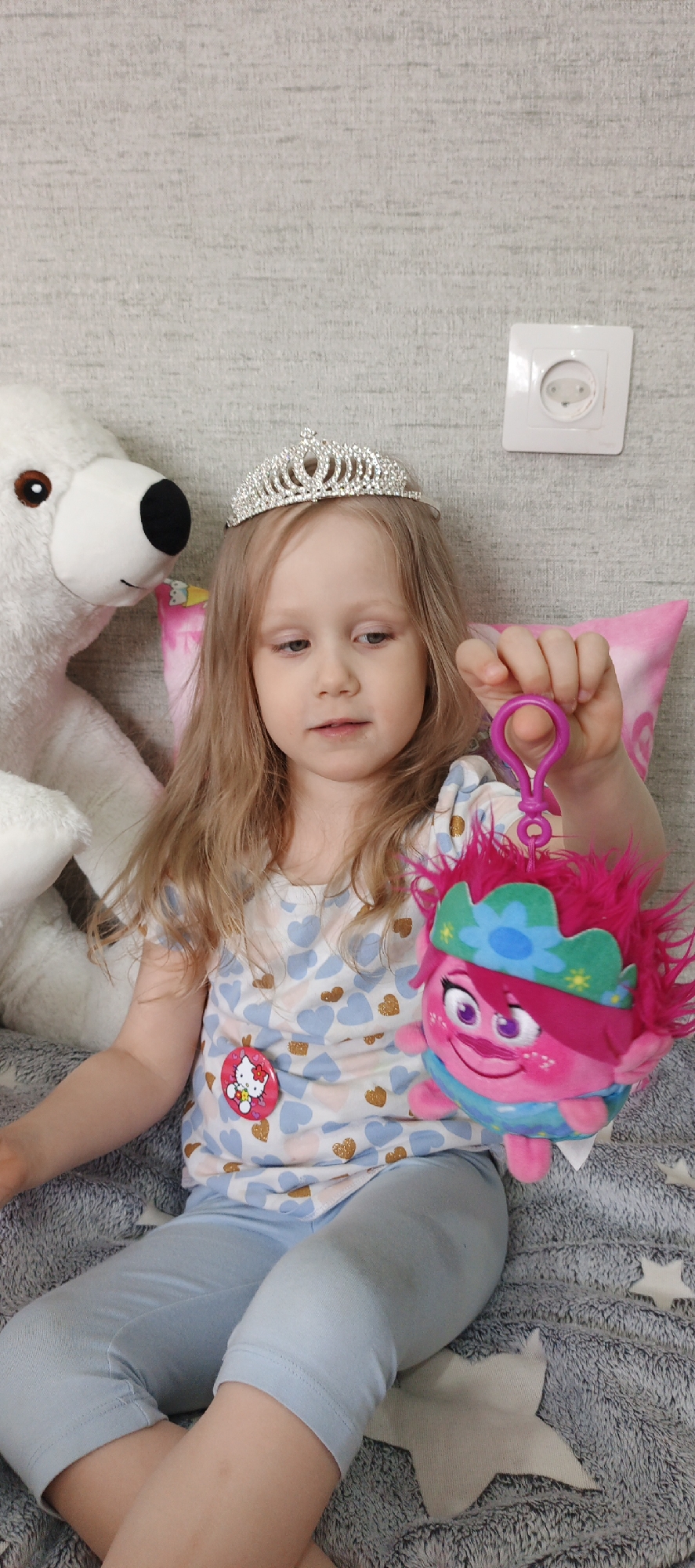 A young girl is seated on a gray blanket with white star shapes on it. She is wearing a silver tiara, a short-sleeved shirt with blue and gold hearts, light blue capri-length leggings, and a Hello Kitty pin. She is holding a pink troll doll with a green and blue crown. A white teddy bear sits behind her to the left, and a pink pillow is behind her to the right. A white electrical outlet is visible on the wall behind her. The location is Chaikovsky, Russia.