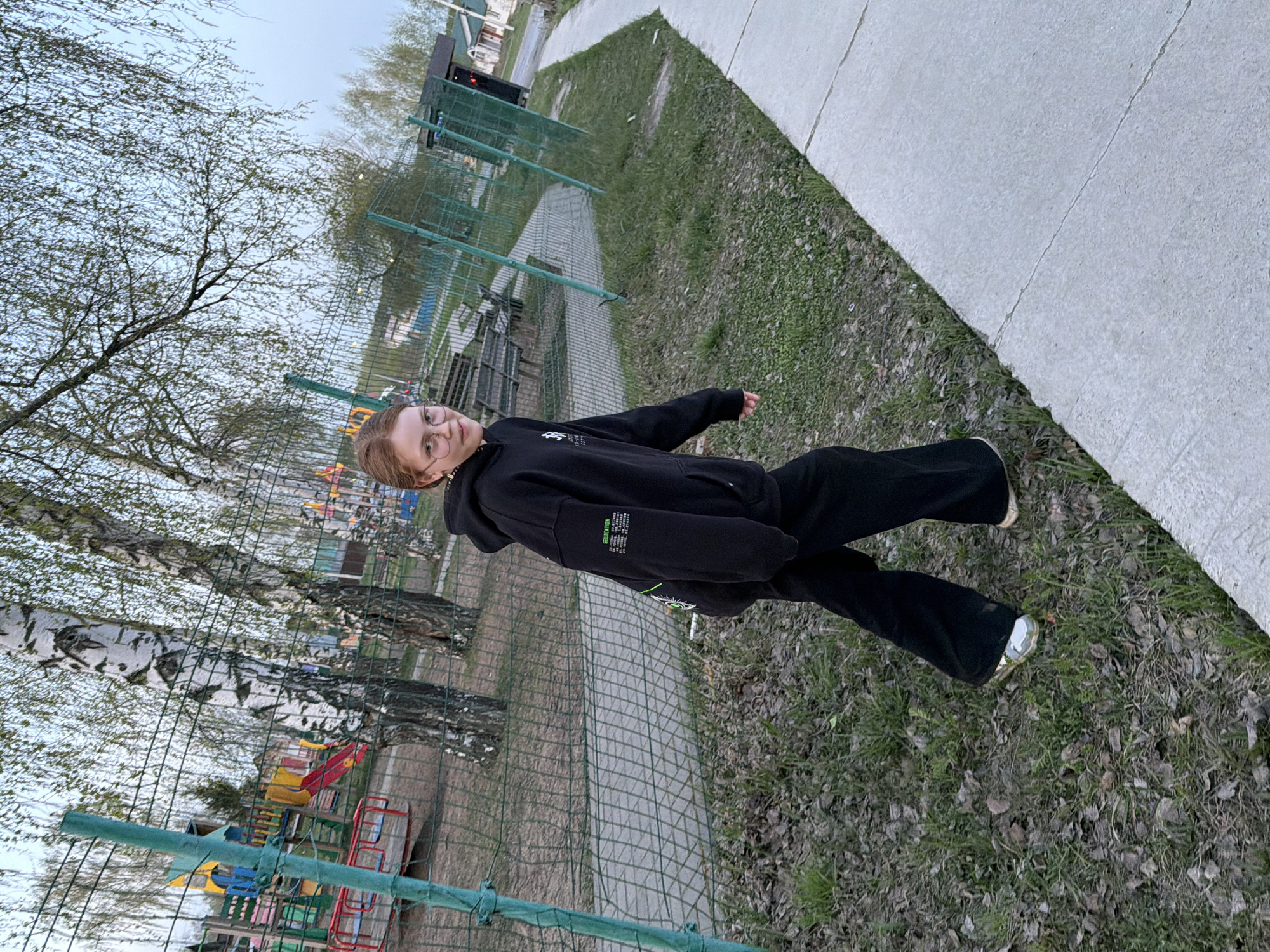 A young person stands outdoors on a grassy area in what appears to be a park or recreational area. The person is wearing a black hooded sweatshirt and black pants, with metallic-colored footwear. To the left, a playground structure is visible behind a chain-link fence, indicating a recreational area. A line of birch trees is also present. The area transitions from grass to a paved path. The image was taken in Chaikovsky, Russia.
