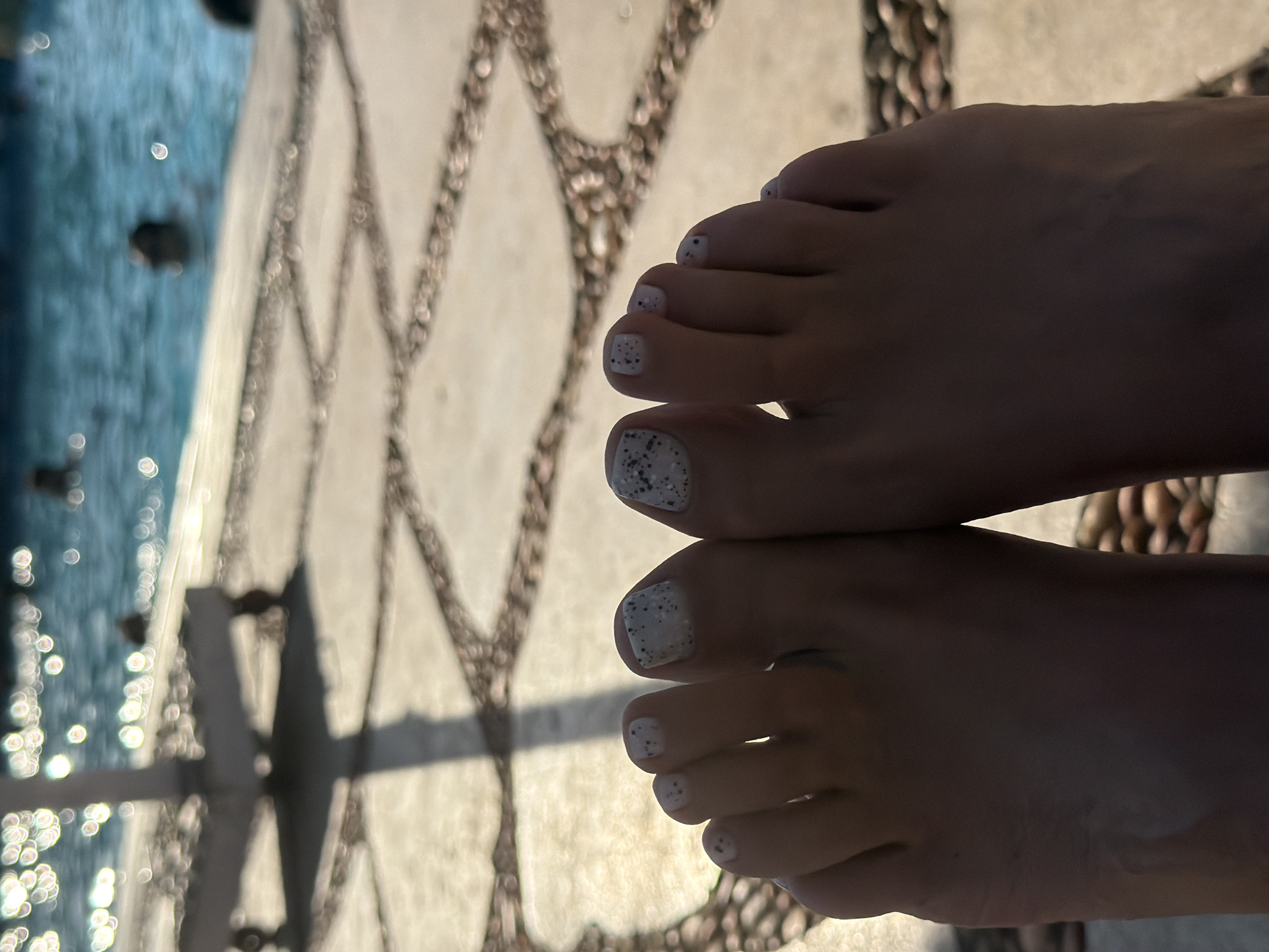 A close-up, low-angle shot features a pair of feet, viewed from the toes up, with a light-colored speckled nail polish. The feet are resting on a paved pathway with a textured, geometric pattern made of light stone tiles and darker, rounded pebbles. In the background, the shimmering blue water of a swimming pool is visible, with blurred figures of people in the water and sunlight reflecting brightly on the surface. A portion of a white poolside table or lounge chair is also visible in the upper left quadrant, casting a shadow. The overall scene suggests a relaxed atmosphere by a pool, likely in a resort area, consistent with locations in Sharm El Sheikh, Egypt.