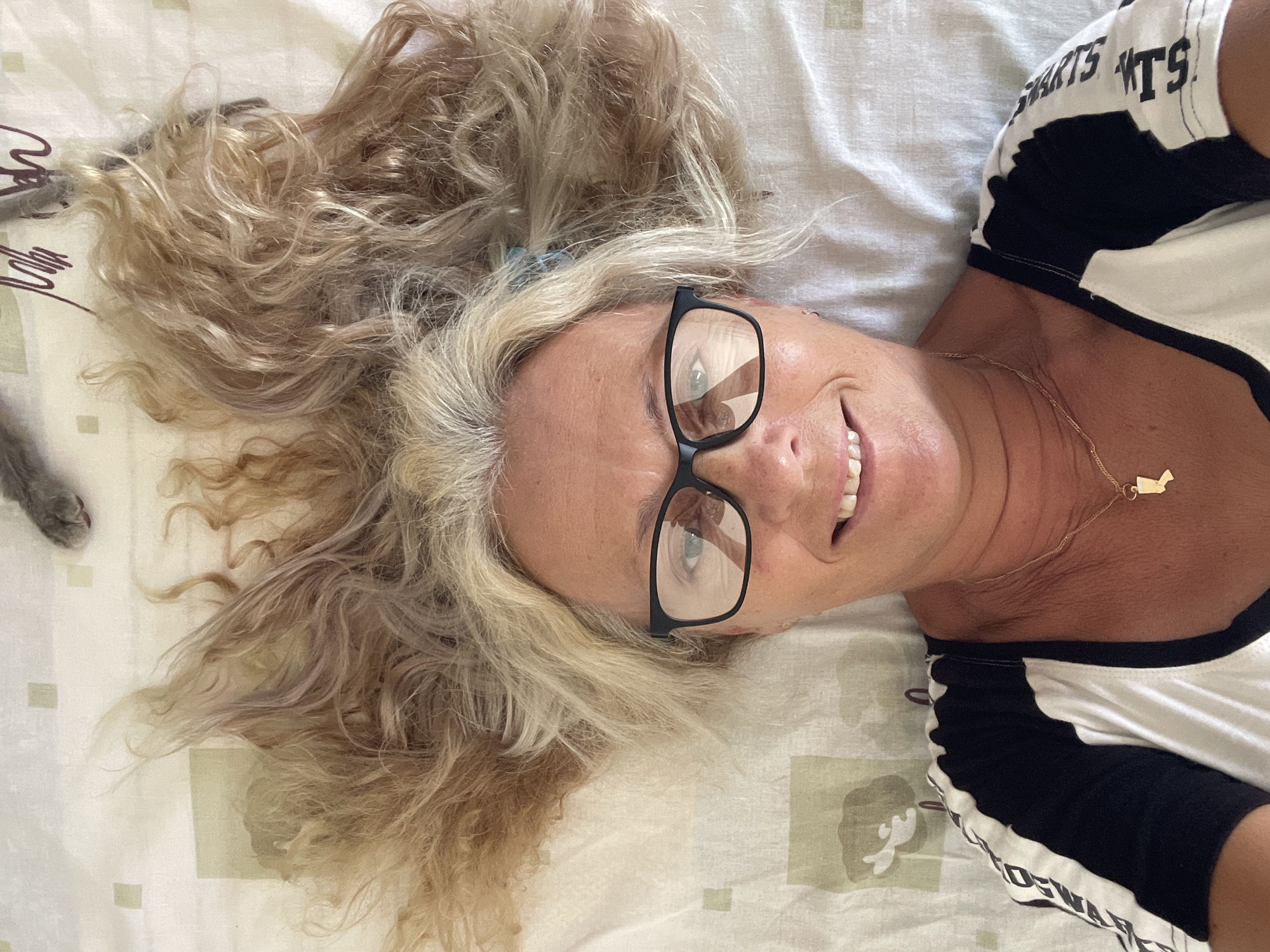 A woman with blonde hair and glasses lies on a bed in Trikomo, Northern Cyprus. Her hair is spread out around her head, resembling butterfly wings. She is wearing a white t-shirt with black stripes on the sleeves. A small gold pendant is visible around her neck. A grey cat's paw is visible in the upper right corner of the image. The bedspread has a subtle green and beige pattern.