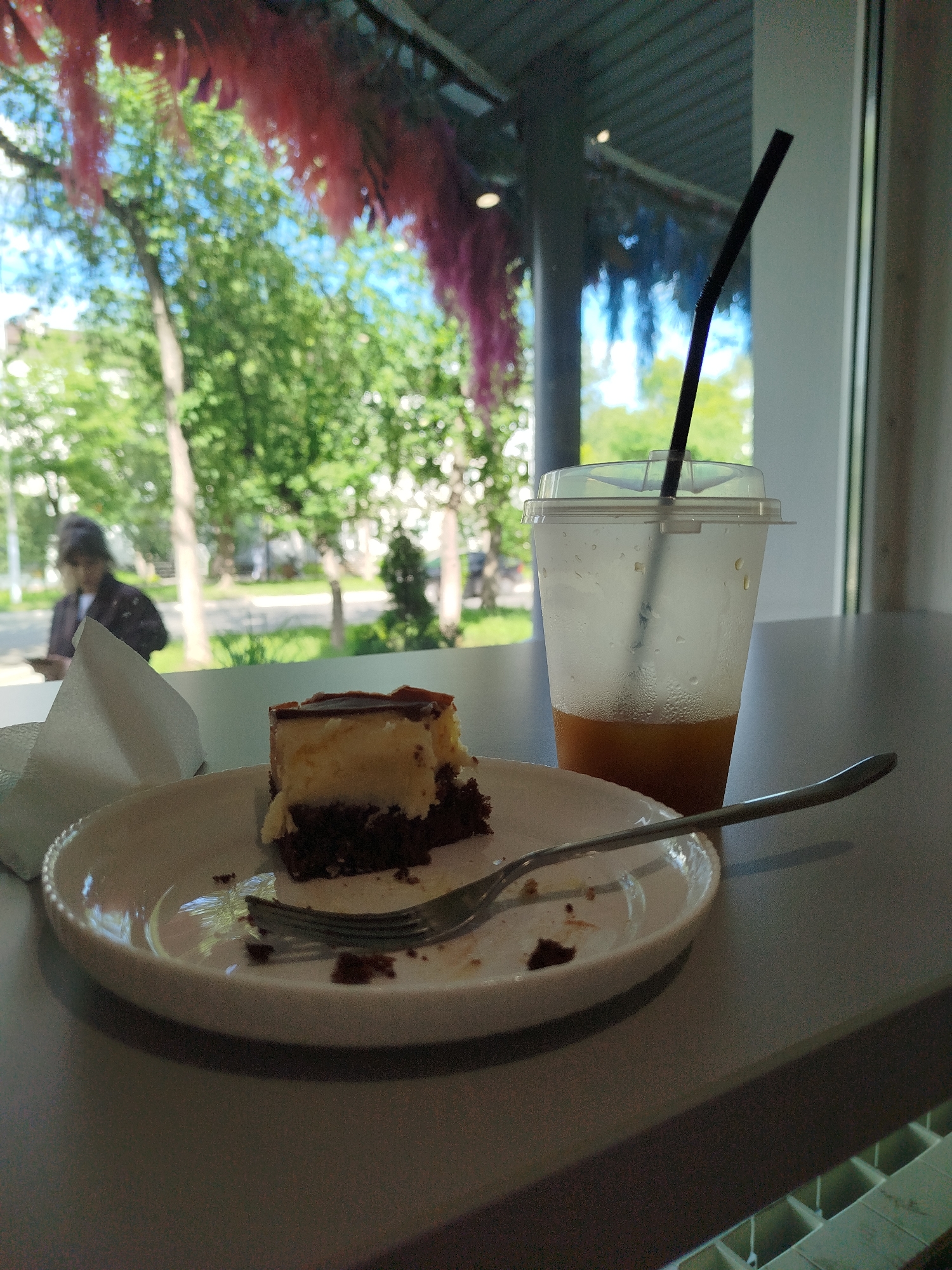 A slice of chocolate cake with a creamy yellow topping sits on a white plate with a fork resting on it. Next to the plate is a clear plastic cup with a black straw, containing an amber-colored liquid. The cup is covered in condensation. In the background, through a window, trees and foliage are visible under a blue sky, with some red and blue decorations hanging from the ceiling. A person wearing a dark jacket and a head covering is blurred in the far left background. The scene appears to be indoors, likely a cafe.