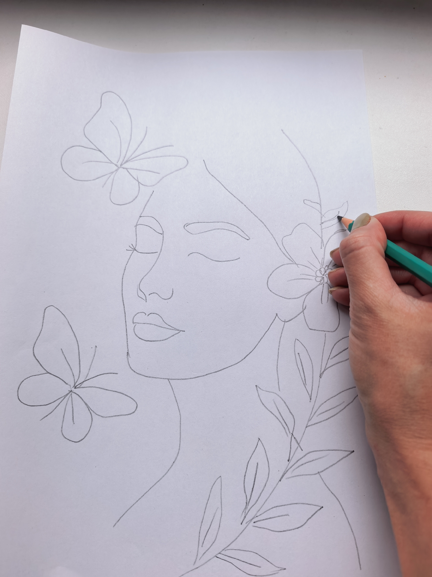 The image shows a pencil sketch of a woman's face with butterflies and flowers drawn around it. A hand holding a green pencil is in the process of drawing on the paper. The sketch is on a white sheet of paper, which is placed on a white surface, likely a windowsill or table. There is no visible indication of the location in the city of Sterlitamak, Russia, or any other geographical context.