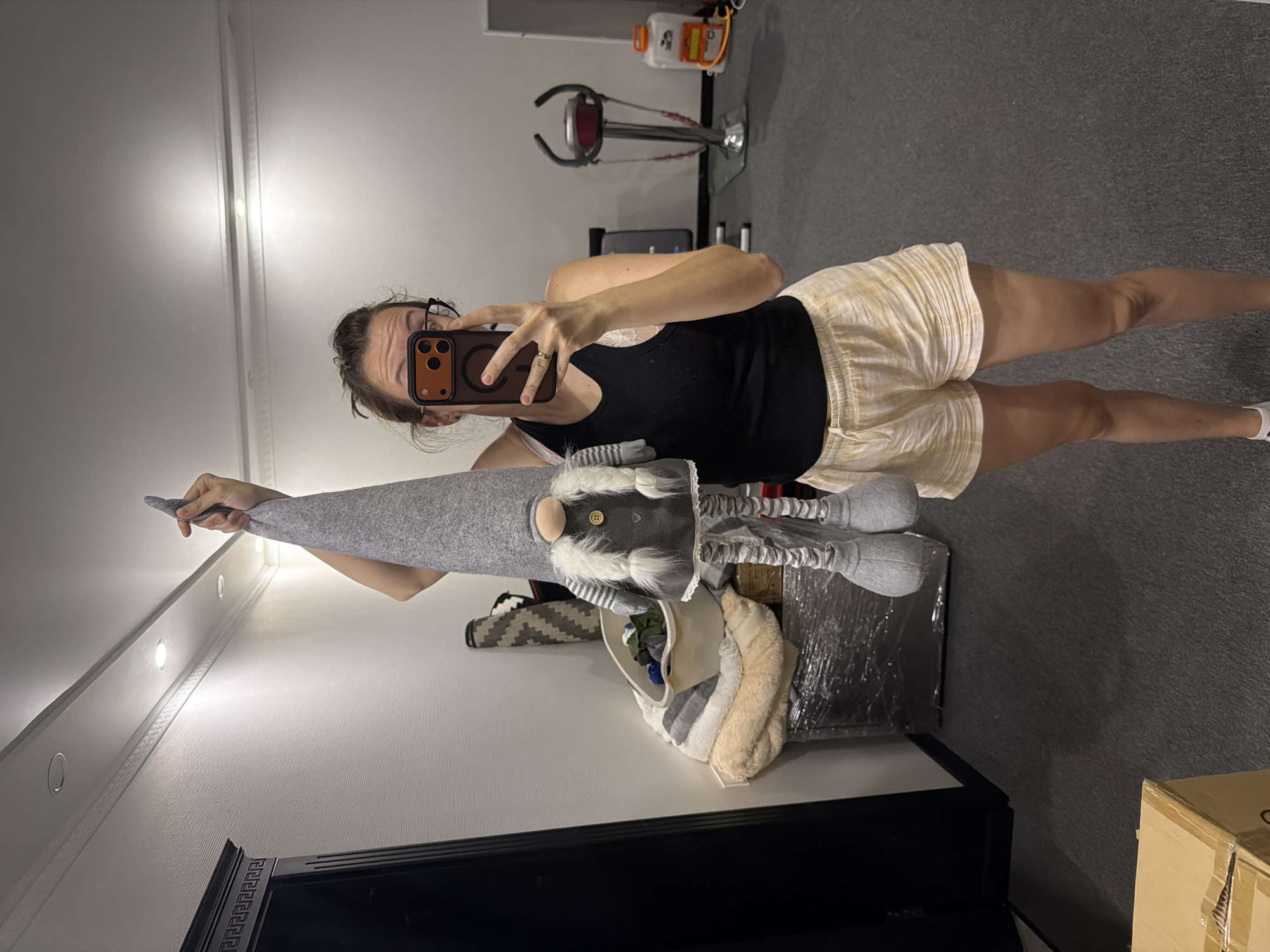 A woman with brown hair pulled back is holding up a tall, grey, plush gnome with a cone-shaped hat. She is wearing a black tank top and light-colored plaid shorts. She is taking a selfie in a room that appears to be a home gym or storage area. In the background, there is a large black wardrobe, a laundry basket filled with clothes, a vibrating exercise machine, and a water cooler. The floor is carpeted. The scene is well-lit by ceiling lights.