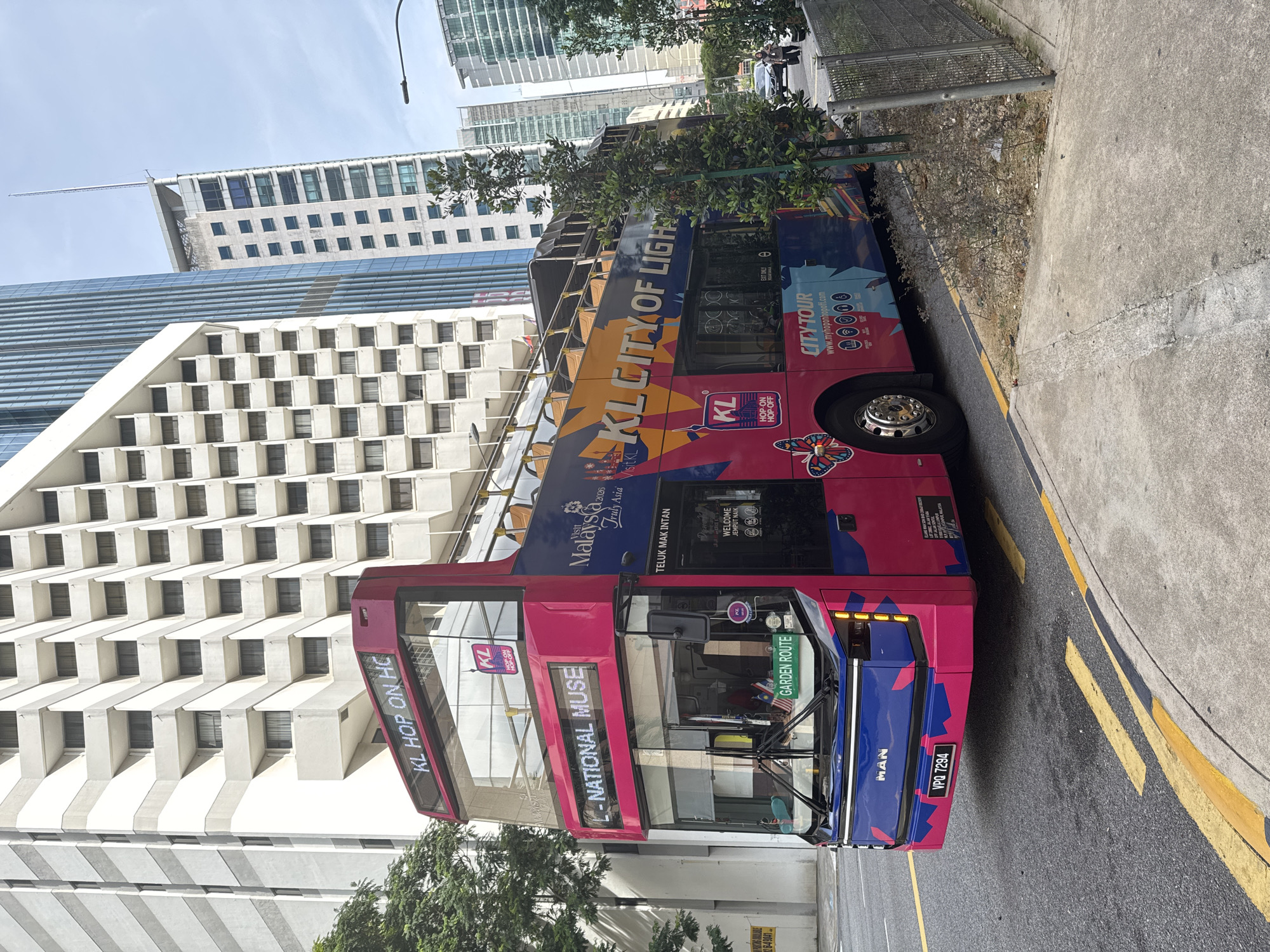A pink and blue double-decker KL Hop On Hop Off city tour bus is parked on the right side of a street in Kuala Lumpur, Malaysia. The bus features advertisements and route information on its side, including "KL CITY OF LIGHTS." In the background, modern high-rise buildings dominate the skyline, including a white hotel with many balconies. The street is paved, with yellow and white lane markings visible. A sidewalk and a metal fence line the right side of the road. The sky is overcast. The license plate of the bus is VPQ 7294.