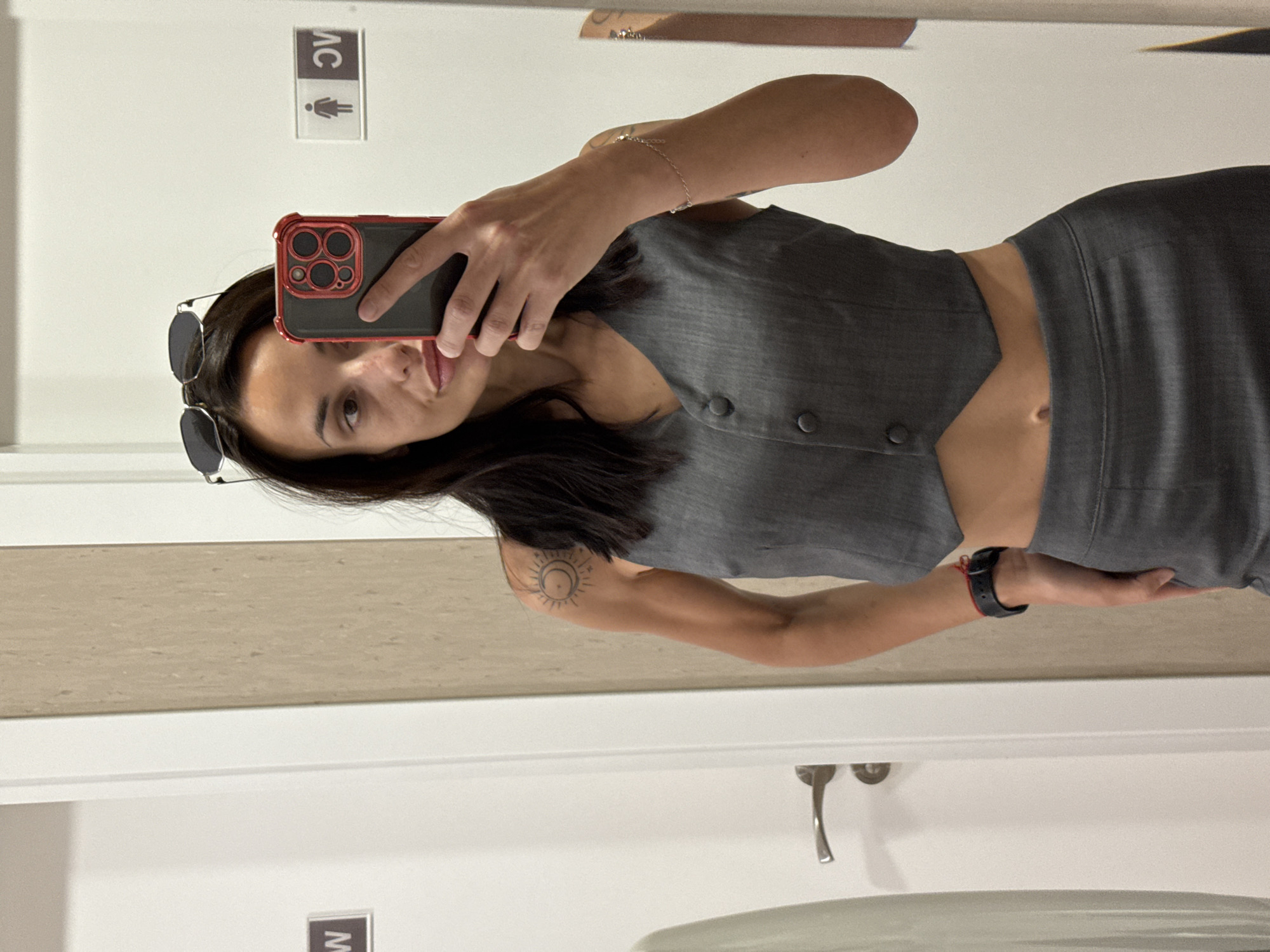 A woman with dark hair, sunglasses on her head, and wearing a gray two-piece outfit is taking a selfie in a bathroom mirror. She is holding a smartphone with a red case in her right hand. The bathroom has a white door with a women's restroom sign and a silver handle. The background includes a beige countertop and a white toilet. The location cannot be determined to be Pleven, Bulgaria from the provided image.