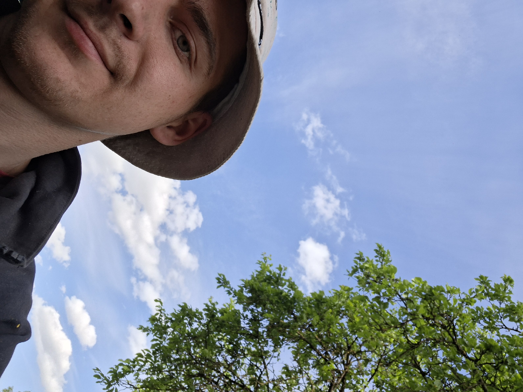A selfie taken from a low angle shows a man's face, wearing a light brown hat, occupying the upper left portion of the frame. His gaze is directed slightly upwards. The right and lower portions of the image feature a bright blue sky with scattered white clouds and the green foliage of a tree. No specific location in Zhabinka, Belarus can be determined from the image.