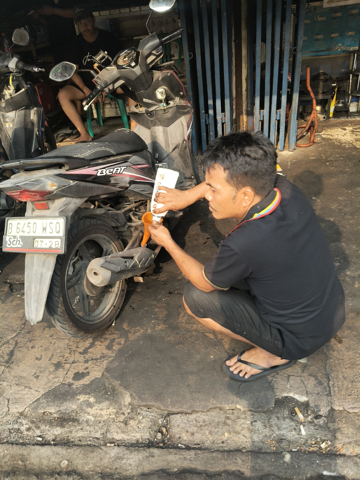 In a South Tangerang, Indonesia, workshop, a man wearing a black shirt and shorts is crouched down next to a motorcycle. He is pouring liquid from a white bottle into an orange funnel that is inserted into the engine of the motorcycle. Another motorcycle is parked to his left, and a person is seated on a chair behind it, partially obscured. The workshop appears to be open-air, with a metal gate in the background and tools and equipment visible. The ground is concrete and appears worn and stained, indicating a working environment. The lighting suggests it is daytime, with shadows cast on the ground. The license plate on the motorcycle reads "B 6450 WSO".