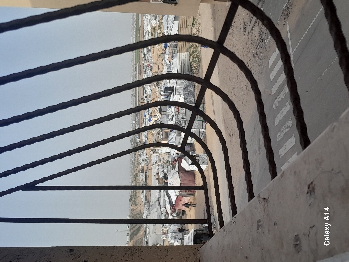 The image presents an elevated view of a scene in Khan Yunis, Palestinian Territory, captured through a dark, rusted metal security grate with multiple vertical bars curving horizontally at the bottom. In the immediate foreground, a worn concrete ledge is visible. Below this, a grey paved road features white pedestrian crossing stripes. Beyond the road, a sprawling encampment occupies a dusty landscape, characterized by numerous densely packed, white and light-colored temporary shelters and tents, some incorporating blue tarps. One individual, dressed in dark clothing, walks on a dirt path within the encampment on the left side of the frame. The horizon line shows indistinct structures and slightly elevated land beneath a hazy, light-colored sky.