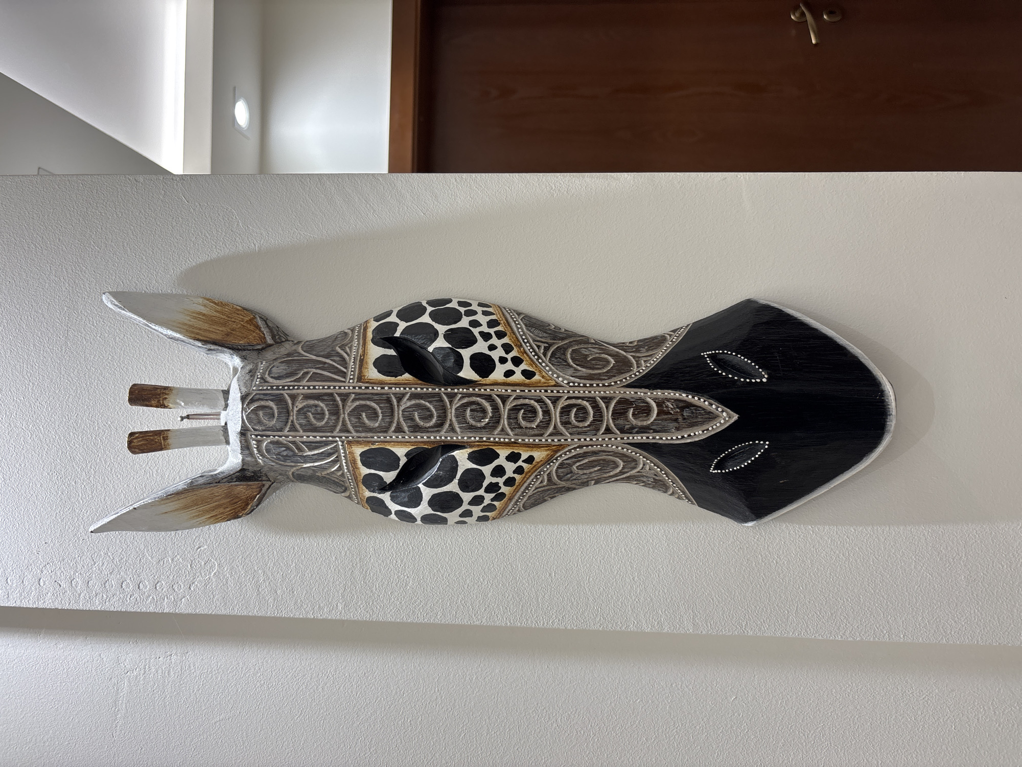 A horizontal close-up shows a decorative wooden giraffe head mask mounted on a light grey wall. The mask is horizontally oriented in the frame, with the giraffe's ears to the left and its snout to the right. The mask's body is decorated with swirled patterns and the head is patterned with black spots on a lighter background. The ears are primarily silver with brown accents, while the snout is black with small white decorative elements. The mask appears to be hand-carved and painted. Above the mask is a dark wood door frame. There is a bright white corner where the wall meets the ceiling in the top left of the image. The location is inferred to be in the city of Plovdiv, Bulgaria, based on external information.