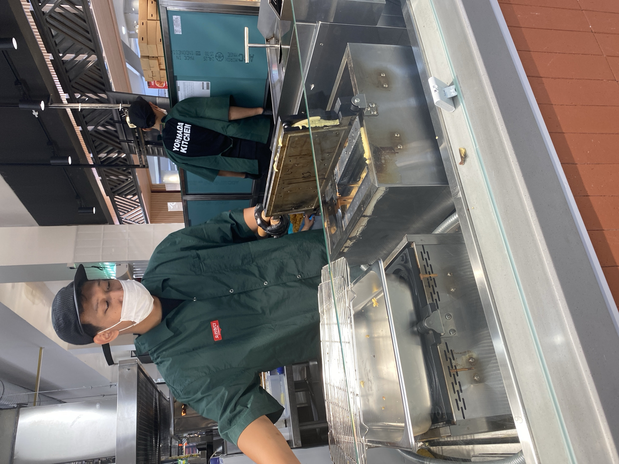 A worker in a dark green uniform, black baseball cap, and white face mask is standing behind a stainless steel counter in what appears to be a food service establishment. He is looking down at a waffle maker which is open, showing batter being cooked. Behind him, another worker wearing a similar uniform and cap is visible, with "YORHADA KITCHEN" printed on the back of his shirt. The environment suggests a clean, modern kitchen or food stall, with stainless steel appliances, tiled walls, and overhead lighting. The scene is captured from a medium distance, focusing on the workers and their activity.