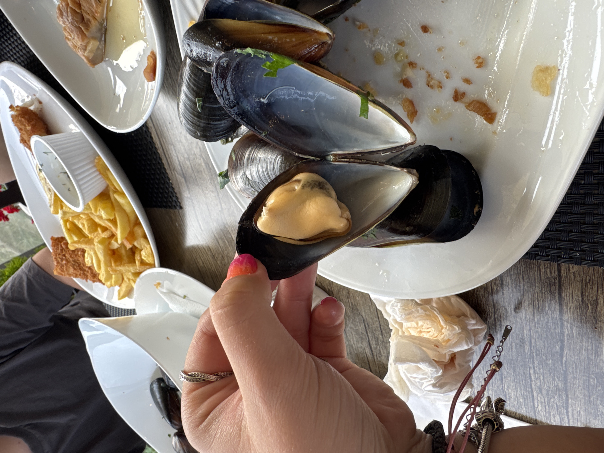 The image depicts a close-up view of a meal at a restaurant, likely located in Sozopol, Bulgaria. A person's hand, adorned with a ring and bright nail polish, is holding a large mussel. The mussel is open, revealing its yellowish-orange flesh. In the background, several other mussels are visible on a white plate, along with some discarded shells and remnants of food.
To the left of the mussels, a white bowl contains more mussels, and a white napkin is crumpled. Further back, a plate holds golden-brown French fries and a small white cup of sauce, suggesting a combination of seafood and fried food. Another white plate in the upper right corner shows a piece of cooked fish in a light-colored liquid. The table surface appears to be made of wood, with a dark, woven placemat visible on the right. The overall impression is a casual dining experience, possibly outdoors given the hint of greenery in the far background and natural lighting.