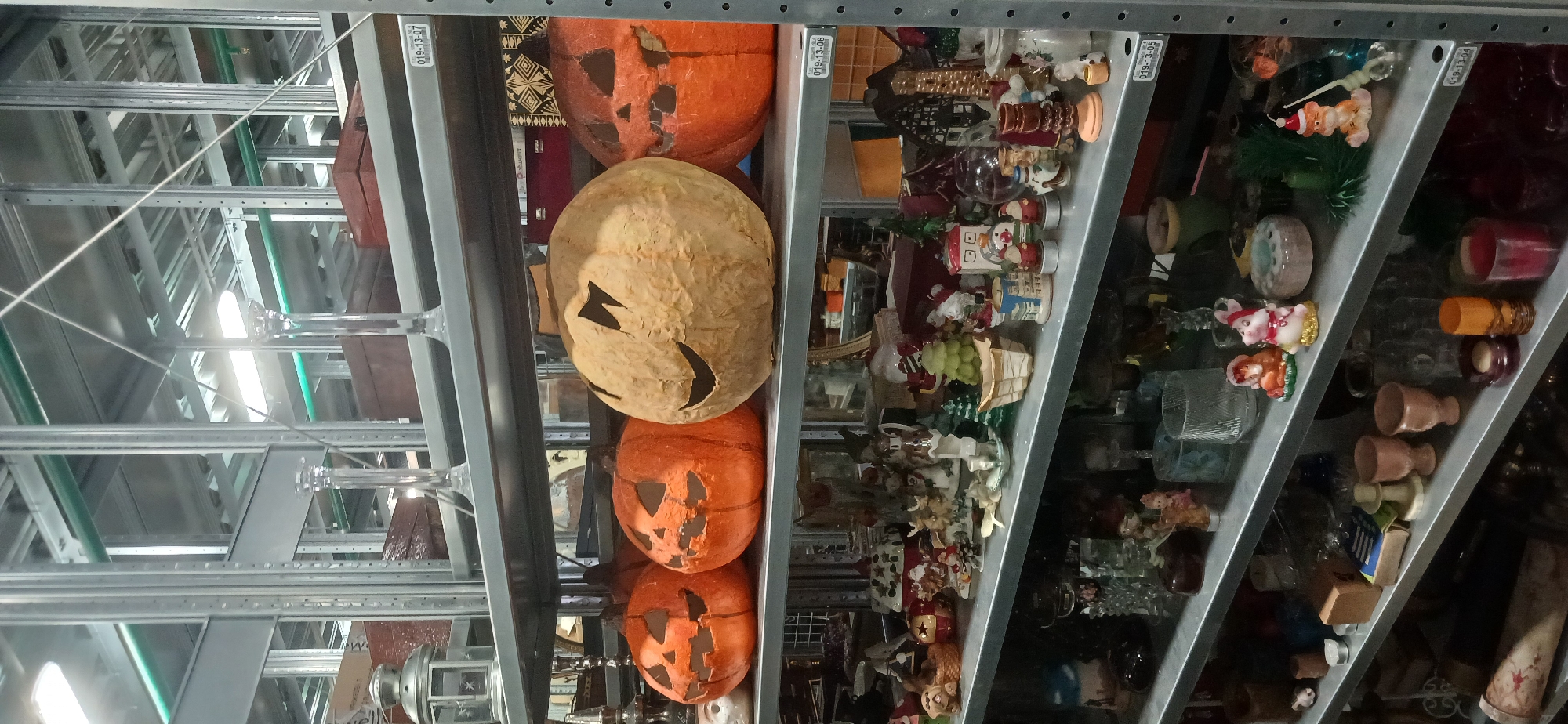 The image displays a series of metal shelving units, likely in a warehouse, thrift store, or backroom of a shop in Moscow, Russia. The shelves are densely packed with a variety of items.
On the left side of the frame, three decorative jack-o'-lanterns are visible. Two are orange, and one has a lighter, tan color, all with carved faces. They appear to be made of a lightweight material such as paper mache or plastic, with some visible wear and tear.
To the right, multiple shelves are filled with a diverse collection of small objects. Many items are holiday-themed, including numerous Christmas figurines such as Santa Clauses, snowmen, and small festive houses or baskets. Alongside these are various pieces of glassware, including cups, vases, and other decorative items. Ceramic figurines, like a pair of mice, small wooden boxes, and miscellaneous trinkets are also present.
The overall setting features industrial metal shelving. Overhead, pipes or conduits (some green) and bright artificial lighting are visible, illuminating the cluttered environment. White labels with inventory codes like "019-13-07," "019-13-06," "019-13-05," and "019-13-04" are affixed to the shelf edges. There are no people, discernible activities, or specific time of day indicated in the image.