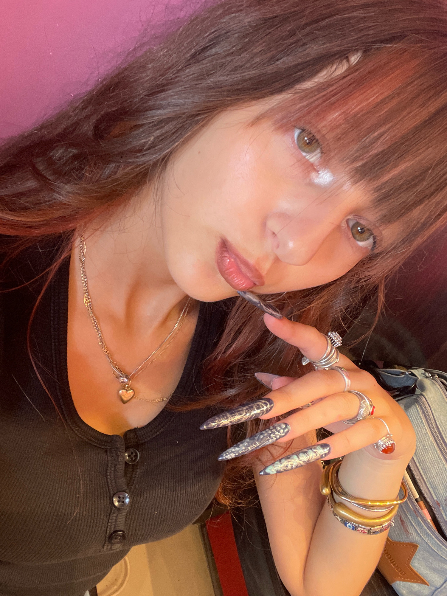 The image shows a close-up of a person's face and upper body, with their right hand prominently displayed. The person has long, dark brown hair with some reddish highlights, and fair skin with visible makeup on their eyes and lips. They are wearing a black, ribbed, button-up top. Around their neck is a silver chain with a heart-shaped pendant. The right hand features long, pointed, dark-colored nails with an intricate design, and is adorned with multiple rings. Several bracelets are visible on the wrist, including gold-toned bangles and a multi-colored beaded bracelet with eye motifs. The background is blurred and appears to be indoors, with a pink wall visible on the left. The image is taken from a high angle, looking down at the subject. The scene does not contain any information that would suggest a specific location in Raanana, Israel.