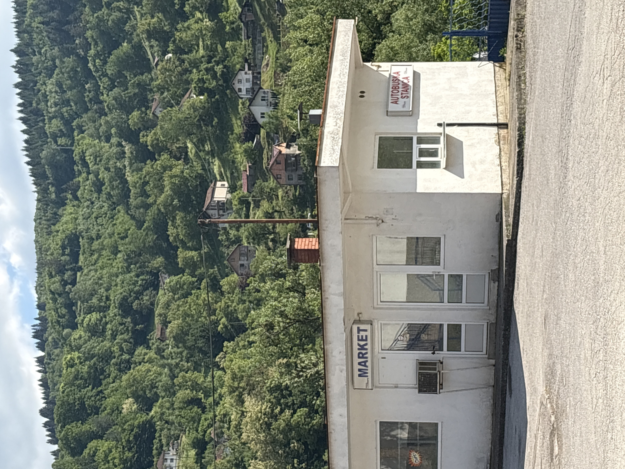 The image captures a scene on the edge of the city of Foča, Bosnia and Herzegovina. The dominant feature is a small, white, two-story building, with signs indicating it serves dual functions: "MARKET" and "AUTOBUSKA STANICA" (Bus Station). The building has windows on both floors and a doorway on the first floor. A small air conditioning unit is attached to the building's exterior.
Adjacent to the building is an asphalt road. In the background, a densely wooded hillside ascends steeply, dotted with houses among the trees. The sky is overcast, providing soft, diffused light. No people are visible in the image. The overall scene suggests a rural or suburban setting on the outskirts of town.