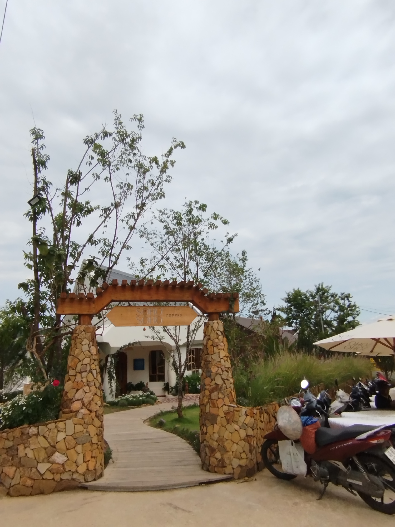 The image depicts the entrance to the "Soho Soho Coffee" shop in Da Lat, Vietnam. The entrance features a rustic stone and wood archway, with the coffee shop sign hanging above the wooden path. The path leads to a white building. A parked motorbike is visible to the right of the entrance, and there are tables and umbrellas further back in the scene. The sky is overcast.