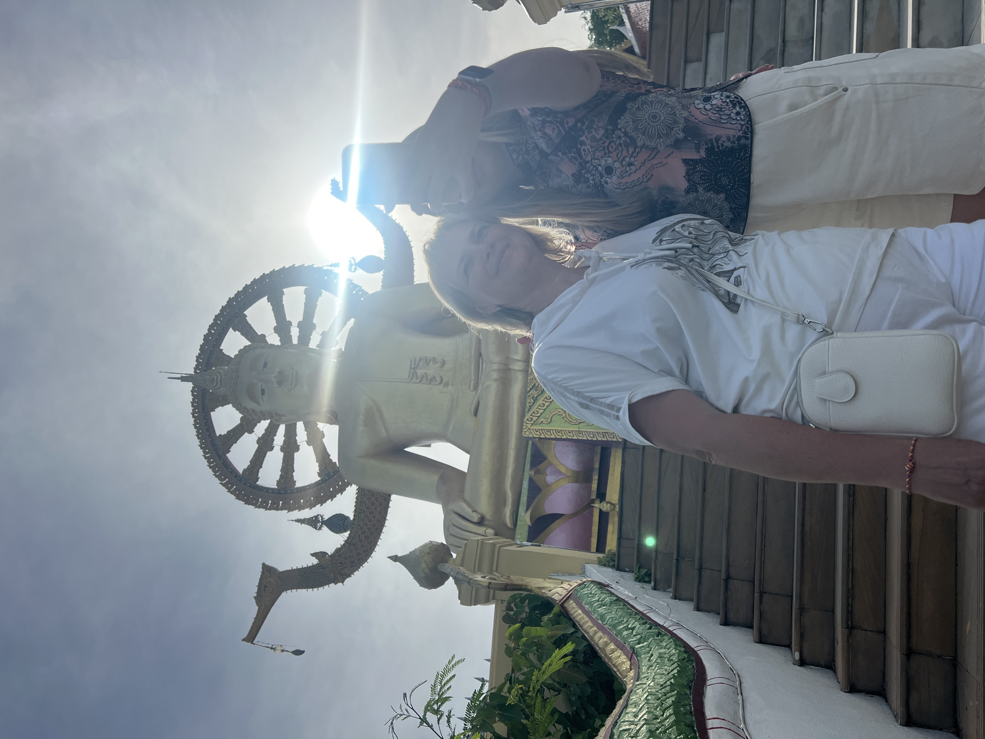 This is a vertical, eye-level, medium shot photograph of two women posing for a selfie in front of a large golden Buddha statue at the Big Buddha Temple in Baan Plai Laem, Thailand. The woman on the left, closer to the camera, is Caucasian with blonde hair and is wearing a white t-shirt and white shorts, and a small white crossbody bag. She is smiling and looking slightly to her left. The woman on the right is holding a smartphone in her right hand, taking the selfie. She has blonde hair and is wearing a black and pink patterned top and white shorts. Her face is partially obscured by the phone. Behind the women, the lower part of a massive, golden Buddha statue is visible. The statue sits on a tiered platform, and a wide staircase with a green Naga railing leads up to it. The sun is bright and positioned in the upper right of the frame, creating lens flare and silhouetting the figures and the top of the Buddha. The sky is bright and hazy. The overall scene suggests a tourist visit to a significant religious site.