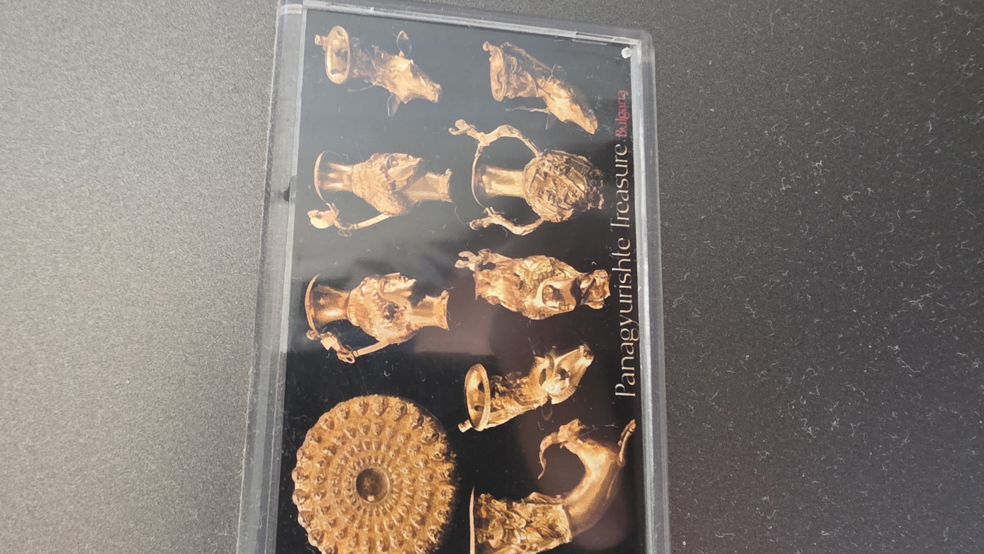 The image captures a clear plastic case, likely for a CD, DVD, or booklet, resting on a textured, mottled grey surface. The cover art inside the case prominently displays nine golden artifacts arranged against a dark, possibly black, background.
These artifacts are part of what is identified by the visible text as the "Panagyurishte Treasure from Bulgaria." The golden objects are intricately designed vessels and decorative pieces, showcasing ancient metalwork. Among them are several rhyta (drinking horns), some crafted in the shape of animal heads such as a stag and a ram, and others resembling human or mythological busts. Two large, ornate amphora-like vessels are also visible, featuring detailed handles and bases. In the bottom left, a circular, shallow dish or phiale with a complex, textured pattern radiates outwards from a central point. Another artifact in the bottom right appears to be the head and neck of an animal, possibly a goat or deer.
The artifacts exhibit a rich, golden hue, suggesting they are made of gold or gold-plated metal. The dark background makes the golden details stand out prominently.
Visible text on the right side of the cover is oriented vertically and reads "Panagyurishte Treasure Bulgaria." The word "Bulgaria" is highlighted in a reddish color, while the other text is in a lighter shade, possibly white or gold. The plastic case itself shows minor reflections and specks of dust. No activity is taking place, as it's a static depiction of historical objects presented on a media cover.