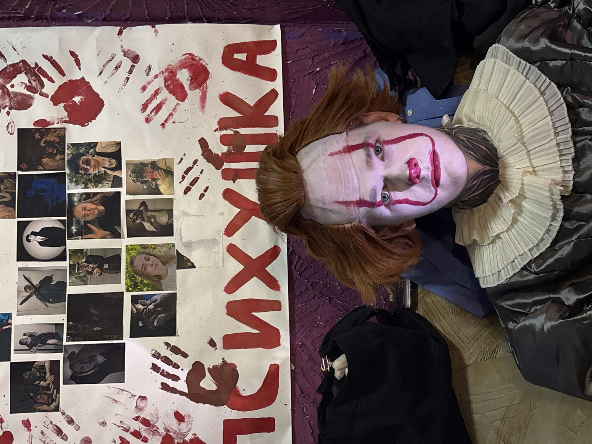 The image depicts a clown doll, resembling Pennywise from the "It" franchise, lying next to a poster with the Cyrillic word "Психушка", which translates to "Psychiatric Hospital" in English. The poster is decorated with red paint handprints and a collage of black-and-white photographs. The doll is wearing a clown costume with a ruffled collar. It is lying on a floor with a patterned rug. The location in Saratov, Russia is unconfirmed.