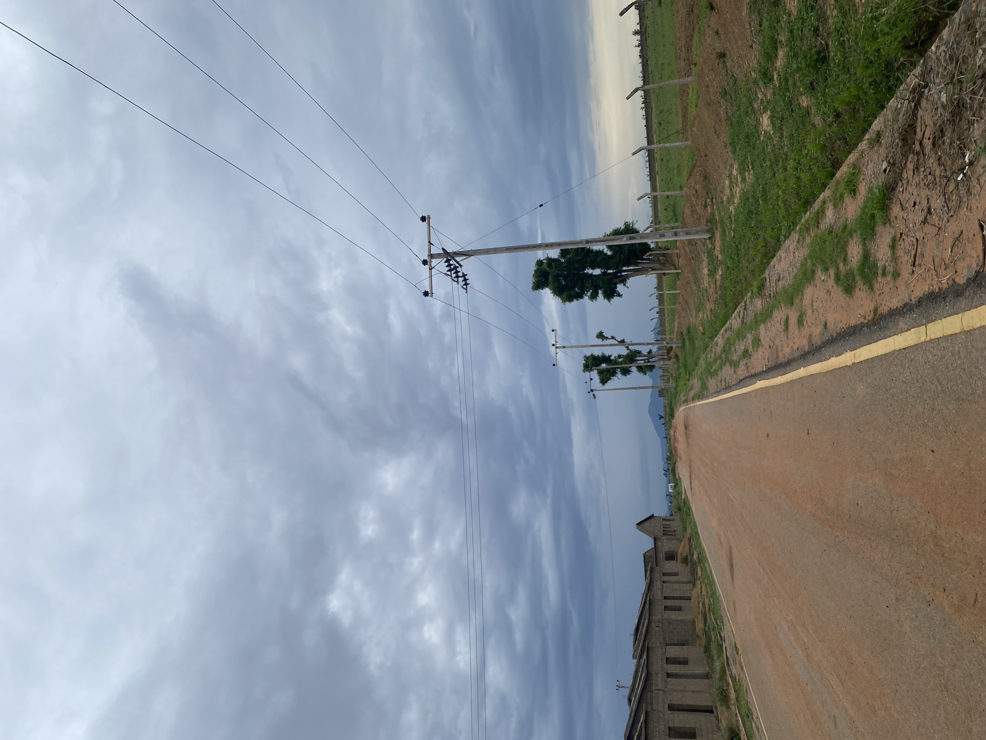 The image shows a dirt road in Dumga, Nigeria, with unfinished buildings on the left and a fenced field with grass on the right. Several utility poles with power lines run along the road. The sky is overcast with dark clouds. The scene appears to be a developing area, with construction in progress and natural elements present.