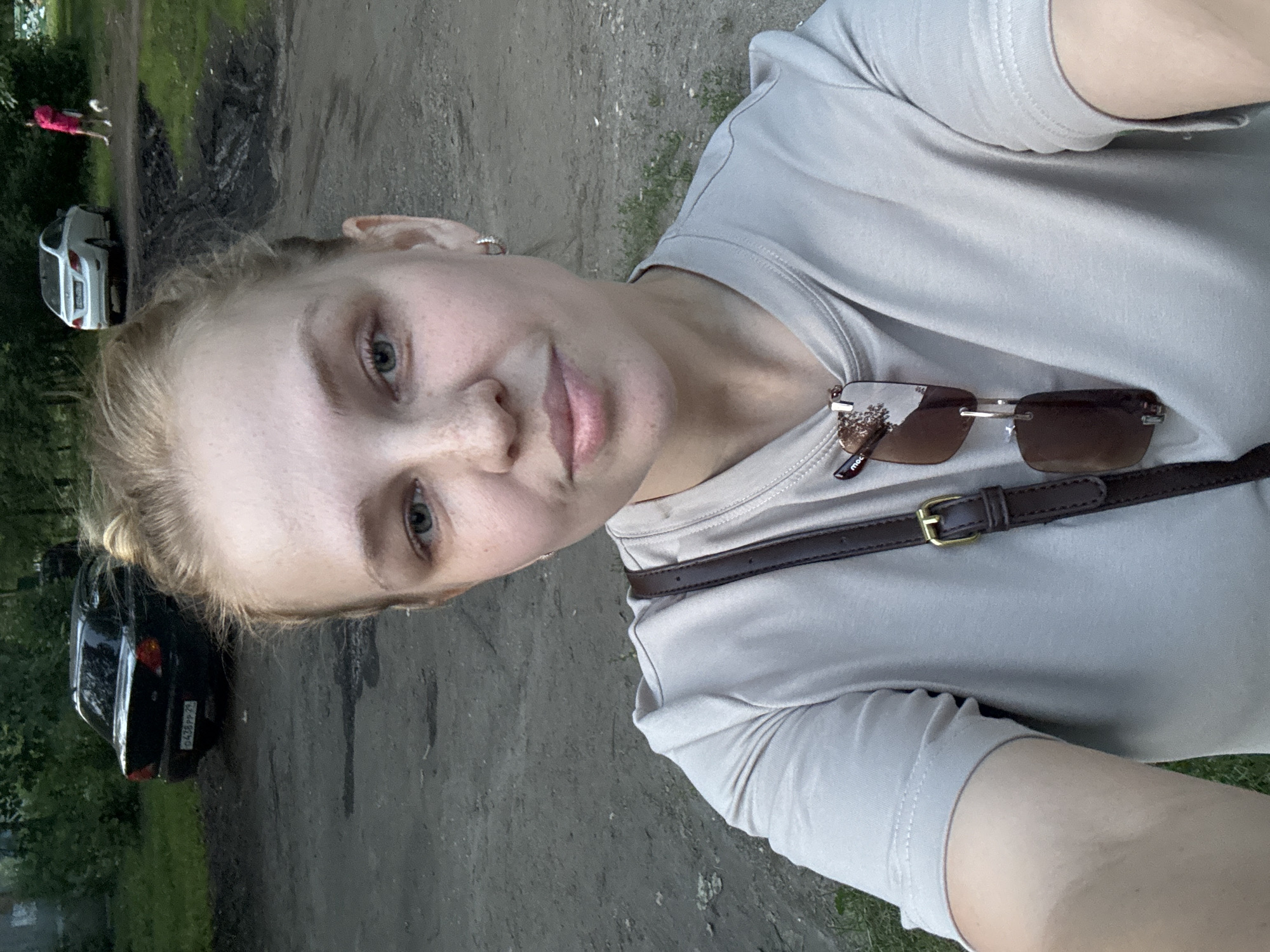 A woman is taking a selfie outdoors. She has blonde hair pulled back in a bun and is wearing a light gray t-shirt. Sunglasses are hanging from the neckline of her shirt. A dark brown strap from a bag is visible crossing her chest. In the background, a black sedan is parked on a dirt road. Further in the background, a white sedan is visible, and a person in a bright pink jacket is walking a small white dog. The area appears to be a residential or common outdoor space with trees and overgrown grass.