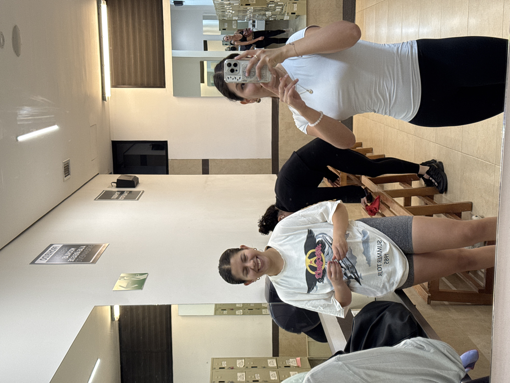 This indoor photograph captures a moment in what appears to be a locker room or changing area, consistent with a location like San Andrés Cholula, Mexico. The image is oriented sideways, appearing rotated 90 degrees clockwise from an upright perspective.
In the foreground, a woman with dark hair pulled back, wearing a white short-sleeved top and black leggings, holds an iPhone with a star-decorated clear case to take a mirror selfie. Her reflection shows her with a neutral to slightly smiling expression, adorned with earrings, necklaces, and bracelets.
To her right, a young girl with dark hair, wearing a white t-shirt with a graphic design (a yellow and red circular logo with wings) and grey shorts, smiles broadly towards the camera. The reflection of her shirt clearly shows the text "SUMMER TOUR 1982". Her hands are clasped in front.
Further in the background, another individual is partially visible, bending over wooden benches, dressed in black activewear and black sneakers with pink accents. In the distant reflection within the large mirrors, two more figures in athletic wear, including one in a sports bra, can be seen, along with metal lockers that line a wall.
The room features white walls, tiled floors, and multiple wooden benches. It is brightly illuminated by long fluorescent light fixtures. Several rectangular signs are visible on the wall; one directly states "PROHIBIDO" (Forbidden), and another reflected sign includes the word "EVACUACION" (Evacuation) along with an emergency exit symbol. The scene suggests a gym, dance studio, or sports facility.