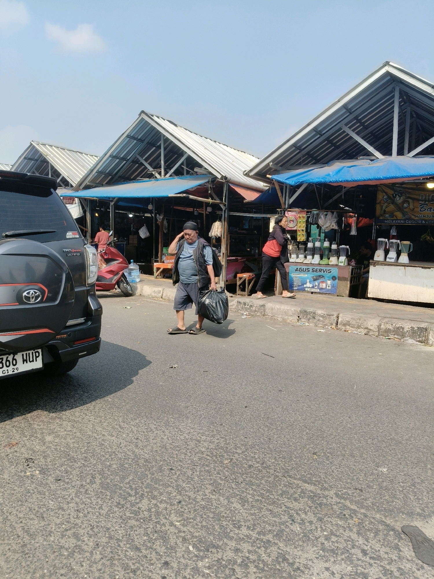 In the city of South Tangerang, Indonesia, a street scene unfolds under a clear sky. In the foreground, the rear of a dark SUV with license plate number 366 WUP is visible on the right side of the road. To the left of the SUV, a red motorcycle is parked, and a large plastic water jug sits on its seat.
A man wearing a dark vest over a grey t-shirt, dark shorts, and sandals is walking on the road, carrying two black plastic bags in his right hand. He appears to be touching his head with his left hand. Further down the road, a woman in a red jacket and black pants is walking on the sidewalk, looking towards the market stalls.
The background features a row of market stalls with peaked, corrugated metal roofs supported by metal frames. Blue tarps are stretched over the fronts of the stalls. One stall displays a sign that reads "AGUS SERVIS" and shows pictures of blenders. Several blenders are lined up on the counter of this stall. Other stalls have various goods displayed, including what appear to be dried goods hanging in some. Another person is partially visible inside one of the market stalls. The market is situated along a paved road with a concrete sidewalk.