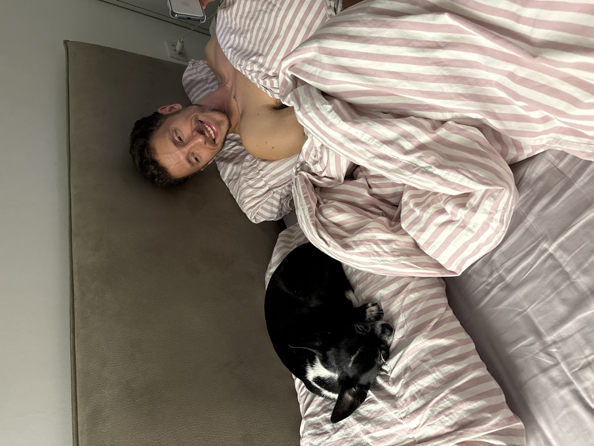 An adult male and a small dog are observed in a bed within an indoor setting, identified as Tel-Aviv, Israel. The male, positioned on the right side of the bed, is partially propped up against a light brown upholstered headboard. He has dark, curly hair and a short beard, is smiling, and is looking towards the camera. He is holding a white smartphone in his right hand, visible at the edge of the frame. A silver chain is around his neck. The dog, black with white markings on its chest and face, is curled up and appears to be sleeping on a pillow to the left of the male. The bed is covered with a light purple fitted sheet. The duvet and pillows feature a light pink and white vertical striped pattern. A white electrical outlet with two plugs is visible on the wall behind the male's head. The overall lighting is dim.