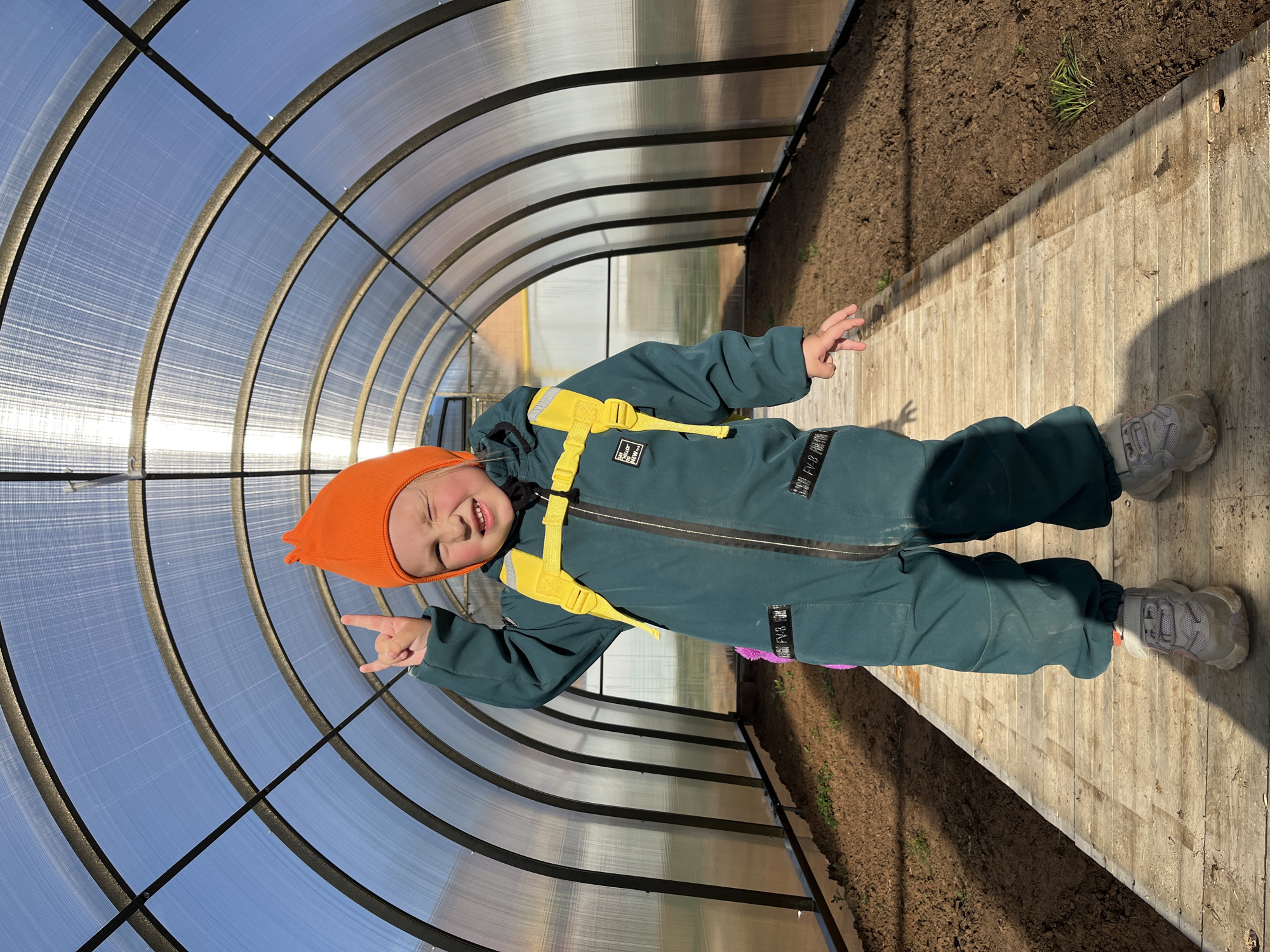 A young child in an orange hat and green jumpsuit with yellow straps is standing in a greenhouse. The greenhouse has a curved plastic roof supported by black metal frames, creating a tunnel-like structure. The child is making a hand gesture. The floor of the greenhouse is a wooden walkway, with soil visible on either side. The location is Chaikovsky, Russia.