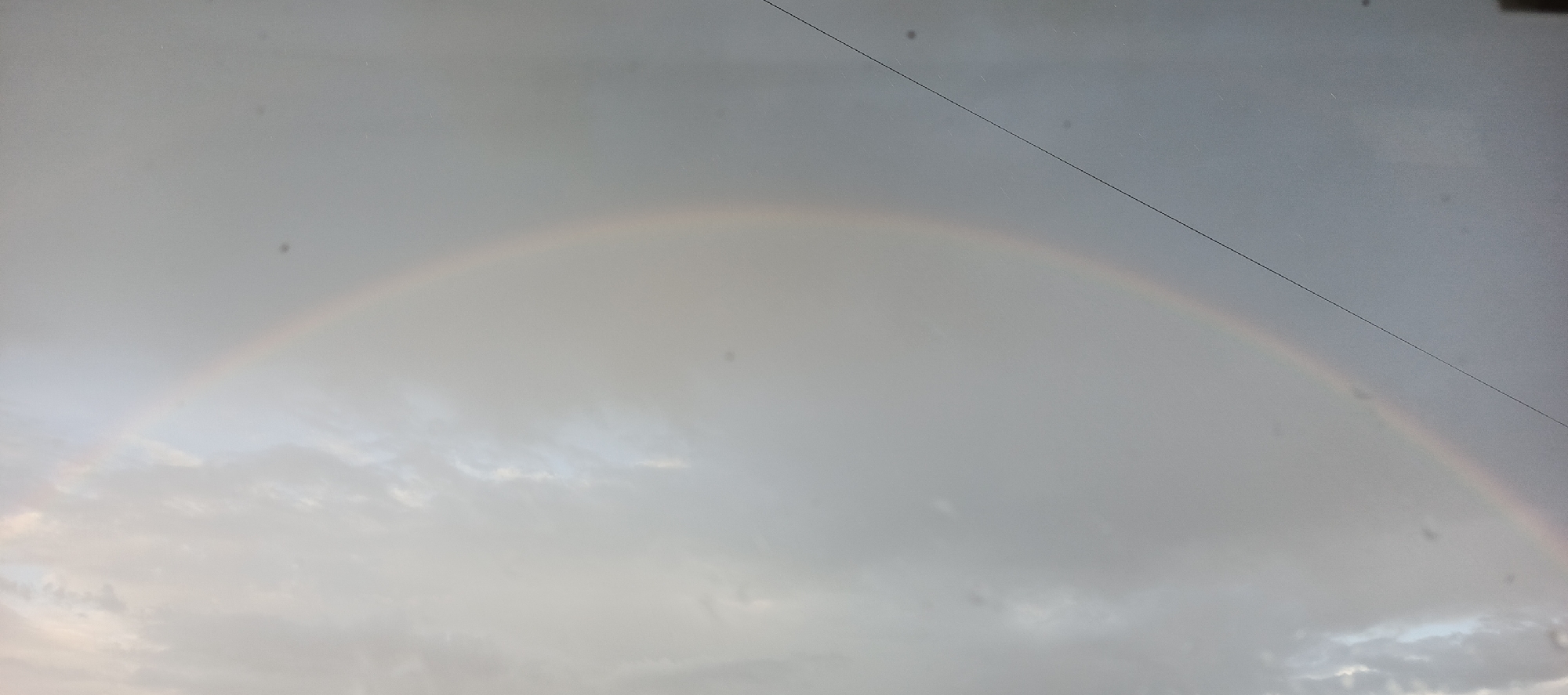 The image shows a faint rainbow arching across a cloudy sky. A power line crosses the upper part of the frame, bisecting the rainbow's arc. The lower portion of the image contains cloud formations. The exact location in Большой Букор, Russia, cannot be determined from the image alone. There are no people or objects in the photo.
