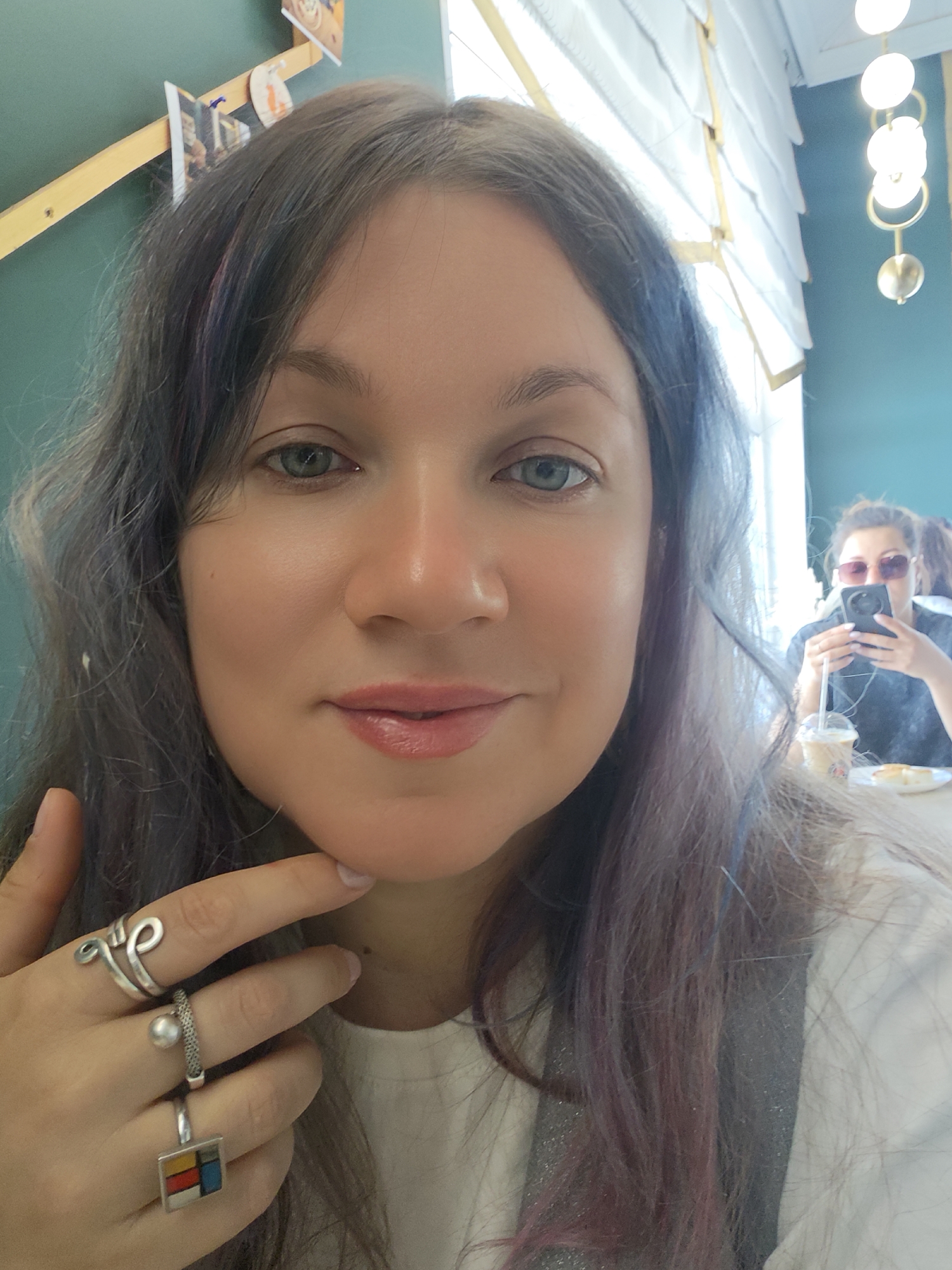 A selfie of a fair-skinned woman with gray-brown hair and dyed strands of purple and blue, wearing multiple silver rings on her left hand. Her left hand is placed under her chin. Behind her, a second woman wearing sunglasses is seated at a table, using a cell phone. A coffee beverage with a straw and a plate with food are on the table in front of her. The background suggests an interior setting with green walls, white window blinds, and a decorative light fixture. Location in the city of Unknown, Unknown.