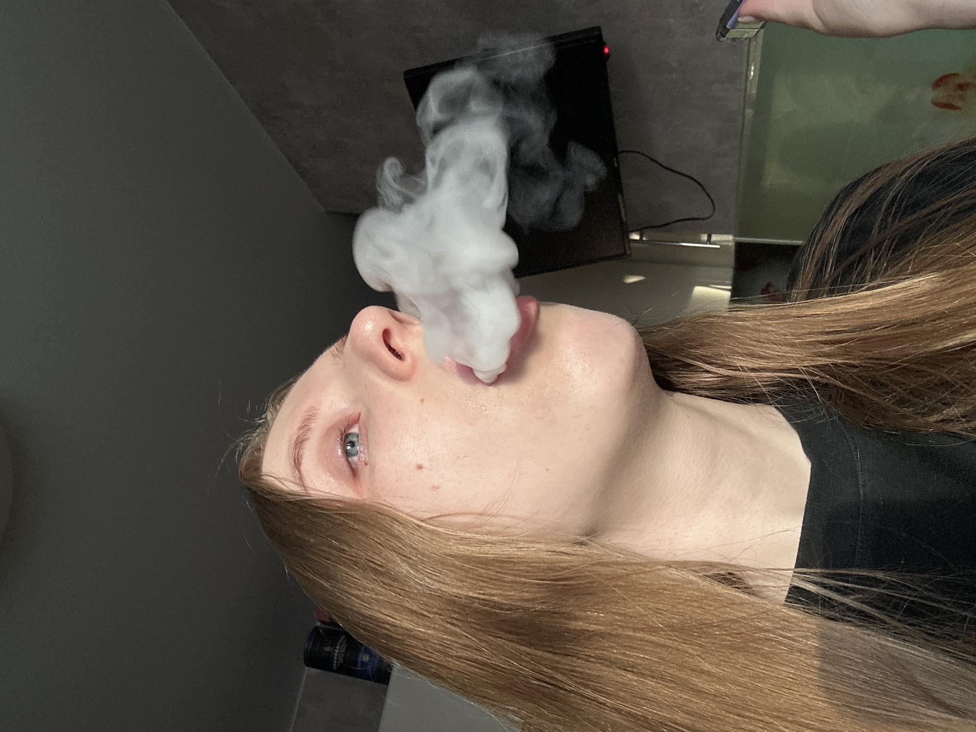 A young adult female, appearing to be in her late teens or early twenties, is depicted exhaling a dense plume of white vapor from her mouth in an indoor setting in Chaikovsky, Russia. Her head is tilted slightly upward and to her left, with her gaze directed towards the upper right. She possesses long, light brown hair, fair skin with visible pores and some freckles, and blue eyes, the left exhibiting slight redness. She is wearing a black garment. In her partially visible right hand, she holds a dark electronic device with a blue illuminated element. The background features a dark shelf to the upper left, holding containers including a dark blue bottle labeled "CHAMBER...". To the right, a wall-mounted black flat-screen television is present, displaying a red standby light at its bottom right corner, indicating it is powered off. Below the television, a large, greenish-tinted glass enclosure, possibly an aquarium, contains indistinct orange-red shapes. The walls are a neutral dark grey or beige, and the ceiling is dark. Lighting emanates from the right, highlighting the subject's right profile and creating shadows on the left.