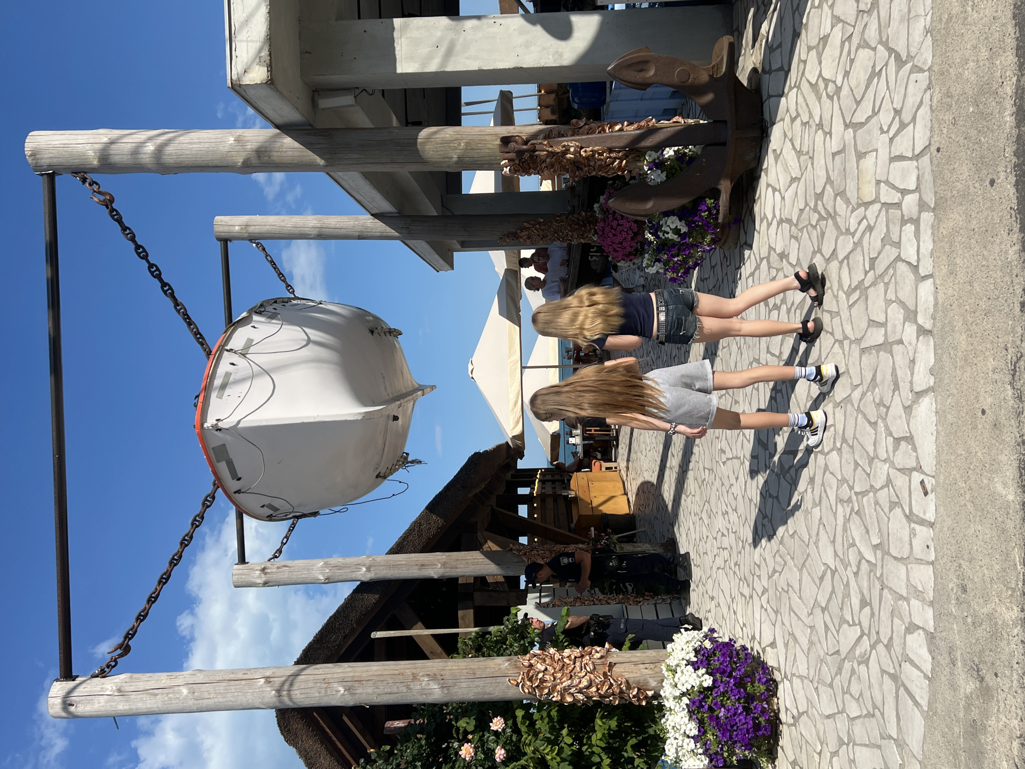 The image depicts a scene in Odesa, Ukraine, on a sunny day. Two young girls with long blonde hair are standing in the foreground, facing away from the camera. The girl on the left is wearing grey shorts and a white t-shirt with yellow stripes on the sleeves, and white sneakers with yellow accents. The girl on the right is wearing denim shorts, a dark blue t-shirt, and black sandals. They are standing on a paved walkway made of irregularly shaped stones.
In the background, a white lifeboat is suspended above the walkway by chains attached to wooden posts. To the left of the walkway is a building with a thatched roof, adorned with a string of what appear to be seashells. Two uniformed police officers are standing near the thatched-roof building. To the right of the walkway is a modern building with a concrete structure. An anchor is positioned next to the concrete building, partially obscured by a large planter filled with purple and white flowers. Several white umbrellas are set up in the distance, suggesting an outdoor seating area. The sky is blue with some scattered white clouds. The overall environment appears to be a coastal area, possibly a harbor or a waterfront promenade.