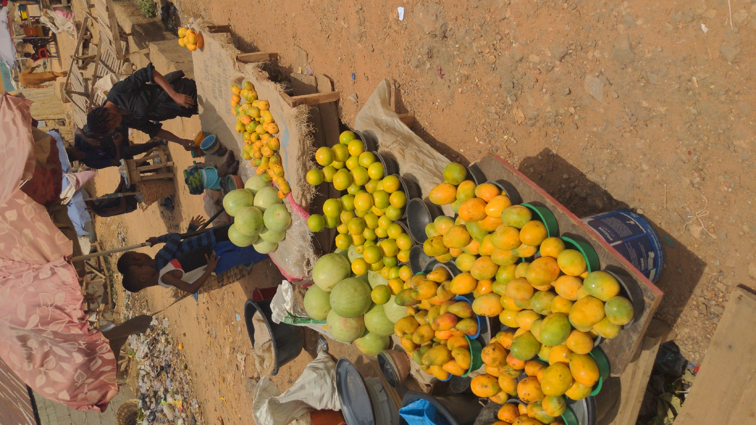 This media file captures a vibrant outdoor market scene in Ado, Nigeria, bustling with activity under bright daylight. The foreground is dominated by large quantities of fresh produce displayed on makeshift wooden stalls and on the ground.
Numerous mangoes, varying in ripeness from green-yellow to bright orange-yellow, are piled high in black and green plastic bowls and buckets across several wooden stands. Alongside these, several large, round green fruits, resembling watermelons or gourds, are arranged on white sacks.
Several individuals are present in the scene. A woman dressed in a black outfit stands behind one of the produce stalls, possibly a vendor attending to customers or organizing her goods. Another adult figure is partially visible near her. In the immediate foreground, two young children are visible; one wears a blue and white striped shirt, and the other a white top, both looking towards the market activity. Other figures are present in the blurred background.
The ground is unpaved, consisting of reddish-brown dirt and gravel, with scattered bits of debris, characteristic of an informal market setting. The bright sunlight casts distinct shadows, indicating it is daytime, likely mid-morning or afternoon. On a blue plastic bucket supporting one of the stalls, white text is partially visible, including "APPLET" and "STORAGE." The overall atmosphere suggests an active, daily market operation.