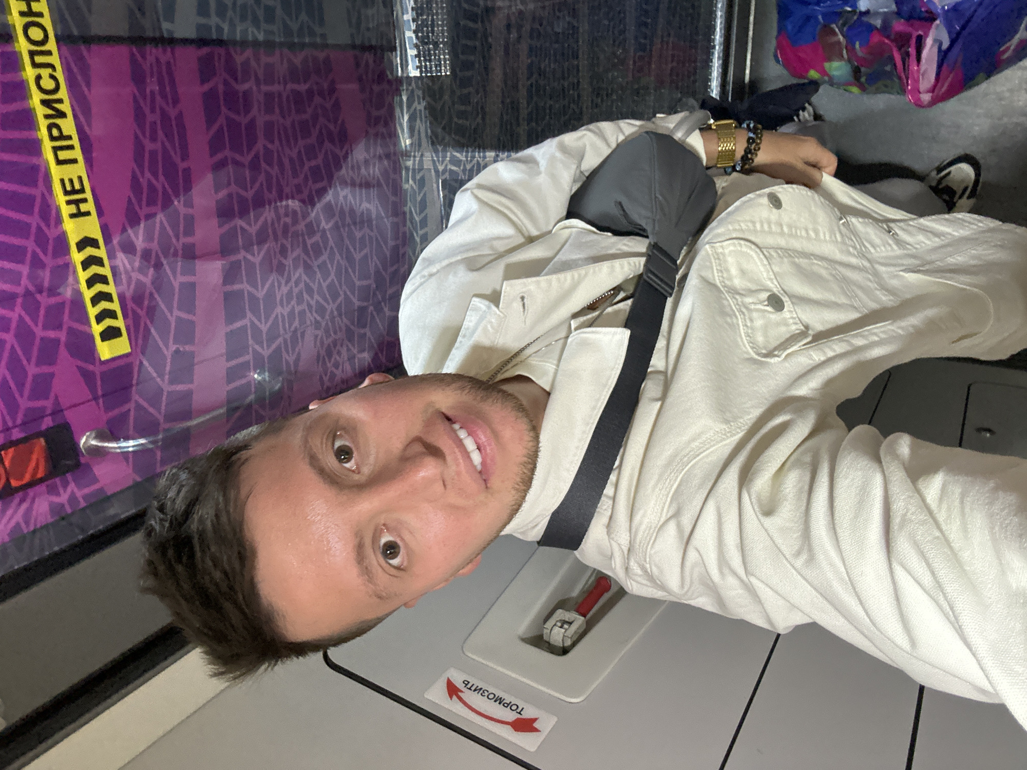 A selfie depicts a man inside what appears to be a passenger train or similar vehicle. He is wearing a light-colored jacket and has a dark-colored shoulder bag. Text in Russian, including the words "НЕ ПРИСЛОН" and "ТОРМОЗИТЬ," is visible on the train's interior. Based on the presence of Cyrillic script and context clues like passenger travel, the location is likely ДСК «Мичуринец», Russia.