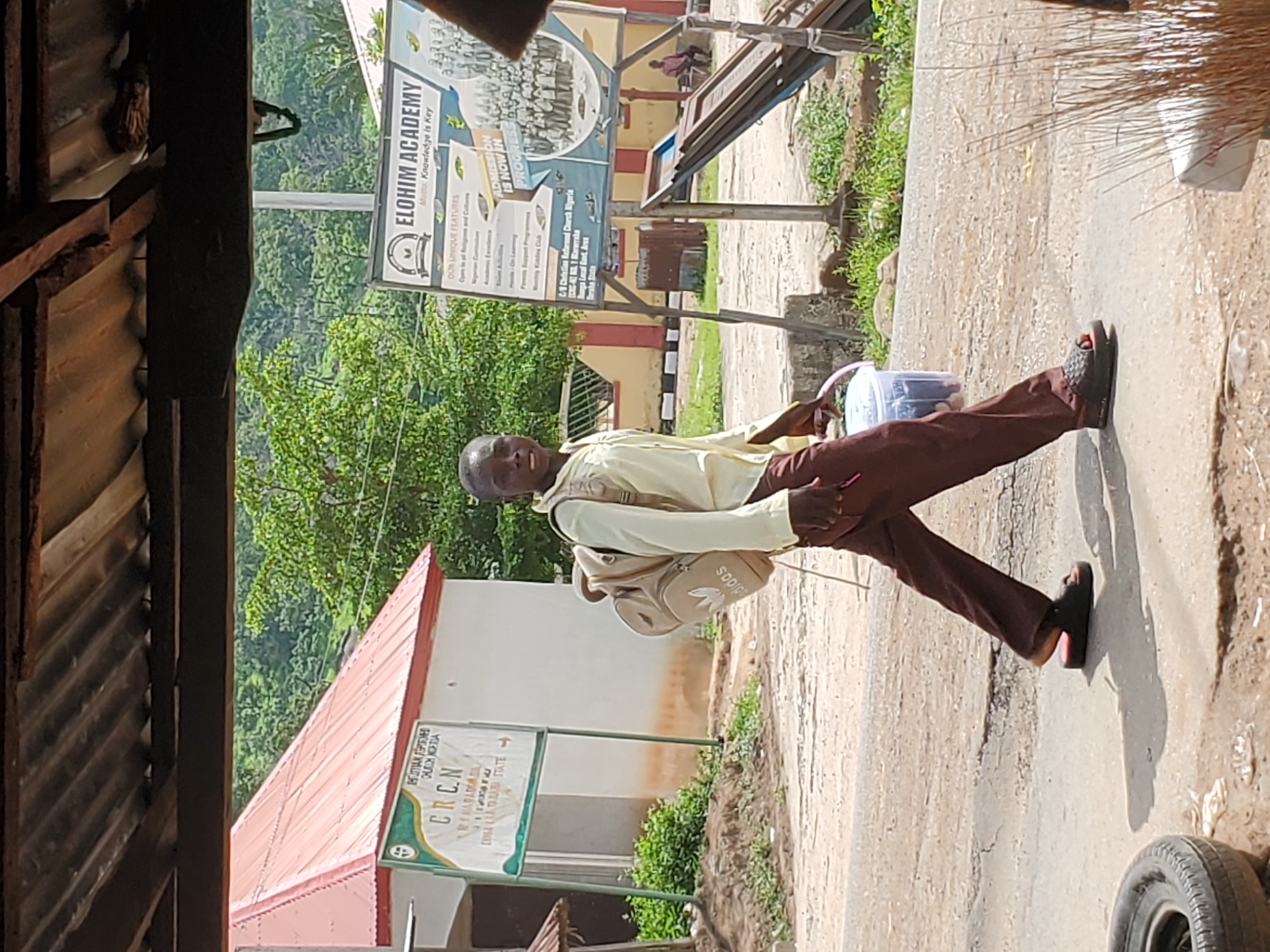 A young Black male walks from left to right across a paved road in Mararaba, Nigeria. He is wearing a light yellow collared shirt, dark brown pants, and dark slide sandals. A light brown backpack with the Adidas logo is on his back, and he carries a clear plastic bucket with a pink handle in his right hand. Behind him, a large sign for "ELOHIM ACADEMY" is visible, listing "Mission: Knowledge is Key" and various school features. To his left, another sign for "C.R.C.N." is mounted on a pole. The background shows trees and vegetation. The ground in front of him is cracked and dusty.