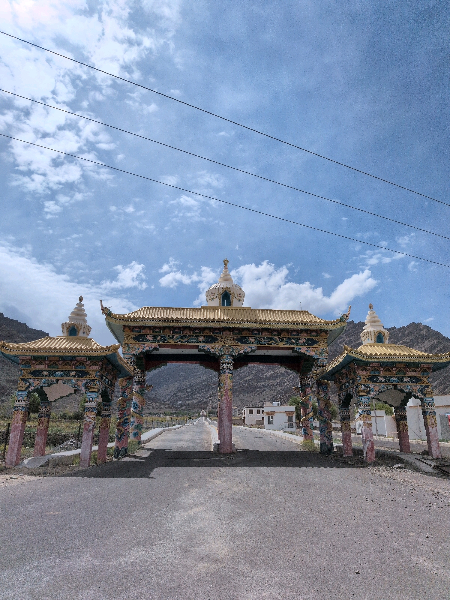 A vibrantly painted Tibetan entrance gate stands prominently in the foreground, its golden roofs and intricately decorated pillars framing a straight, asphalt road that recedes into the distance. The gate is adorned with Buddhist iconography, including stupas on top of its pavilions and colorful murals on its columns. Beyond the gate, the road passes through a sparsely populated area with low-lying buildings and dry, mountainous terrain. The sky above is a bright blue, partially filled with fluffy white clouds, and several black power lines stretch horizontally across the upper portion of the frame. The overall scene suggests a location in a high-altitude, possibly remote, region, consistent with Martselang, India, known for its Tibetan Buddhist culture.