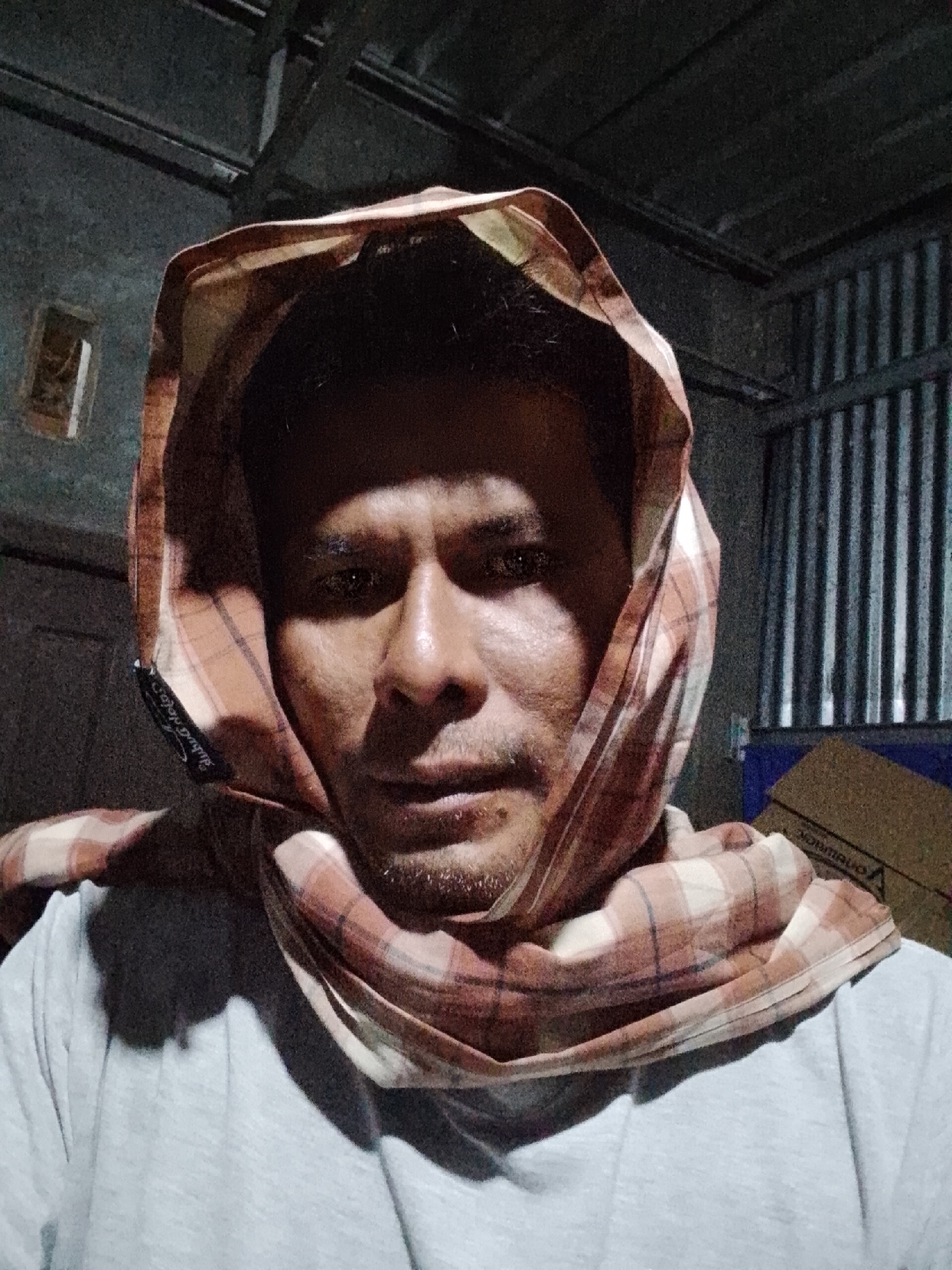 A man is pictured from the chest up, wearing a checkered headscarf over his head and a grey t-shirt. He has a light beard and is looking directly at the camera. The background appears to be an interior space with corrugated metal walls and visible ceiling beams. A cardboard box is visible in the lower right corner. The image appears to be taken in a dimly lit environment, possibly a workshop or storage area, located in the city of Padang Dalom, Indonesia.