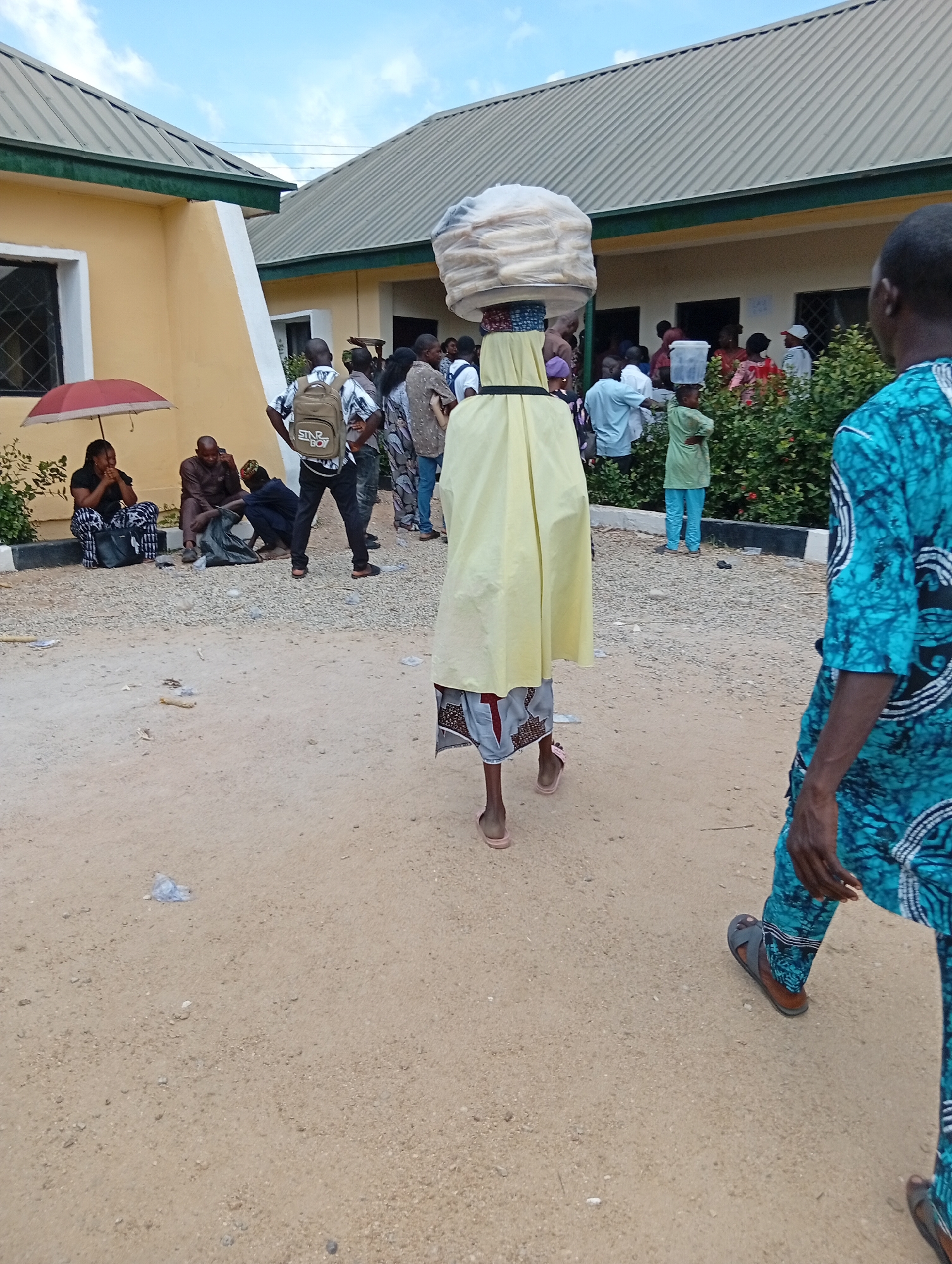 A group of people are gathered outside two yellow buildings with corrugated metal roofs, suggesting a public facility or marketplace in Jalingo, Nigeria. The foreground features a person in a teal and blue patterned robe walking away from the camera, while a young person in a light yellow garment carries a large stack of items wrapped in plastic on their head. Other individuals are visible standing and sitting in the background, some near a red umbrella and others in front of the buildings. The ground is a mixture of sand and gravel. The sky above is partly cloudy.