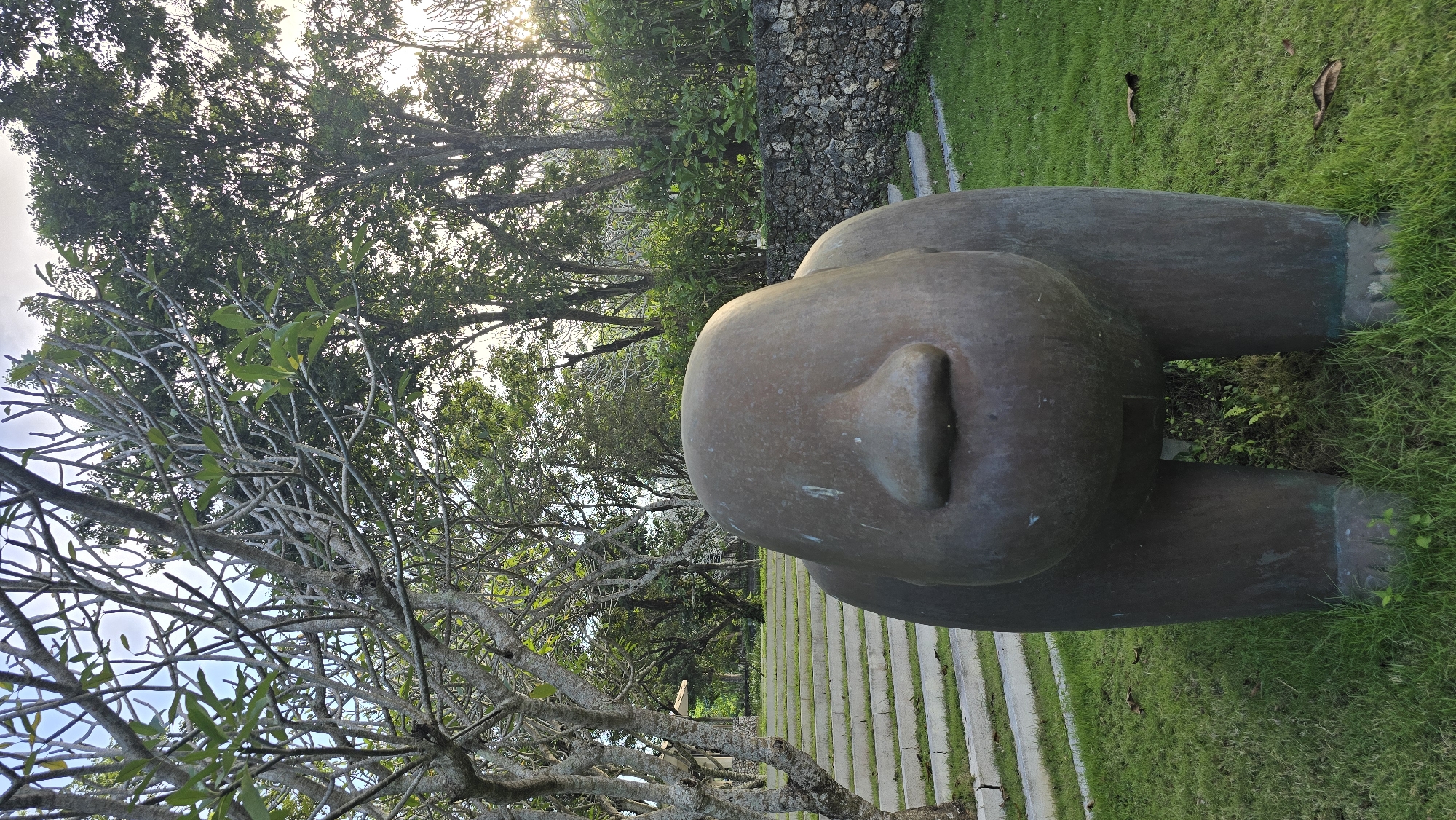 The image shows a bronze sculpture of a person lying on their stomach with their head resting on the ground in a lush outdoor setting. The sculpture is centrally positioned in the frame. The background features a mix of dense tropical vegetation, including trees with light-colored branches in the foreground and taller, leafy trees in the distance. A wall constructed of irregular stones is visible behind the sculpture. A path made of light-colored slabs interspersed with grass leads away from the sculpture. The photo appears to be taken at Kuta Selatan, Indonesia, judging by the location tag and typical landscape features. There is no other visible evidence of people or other activities.