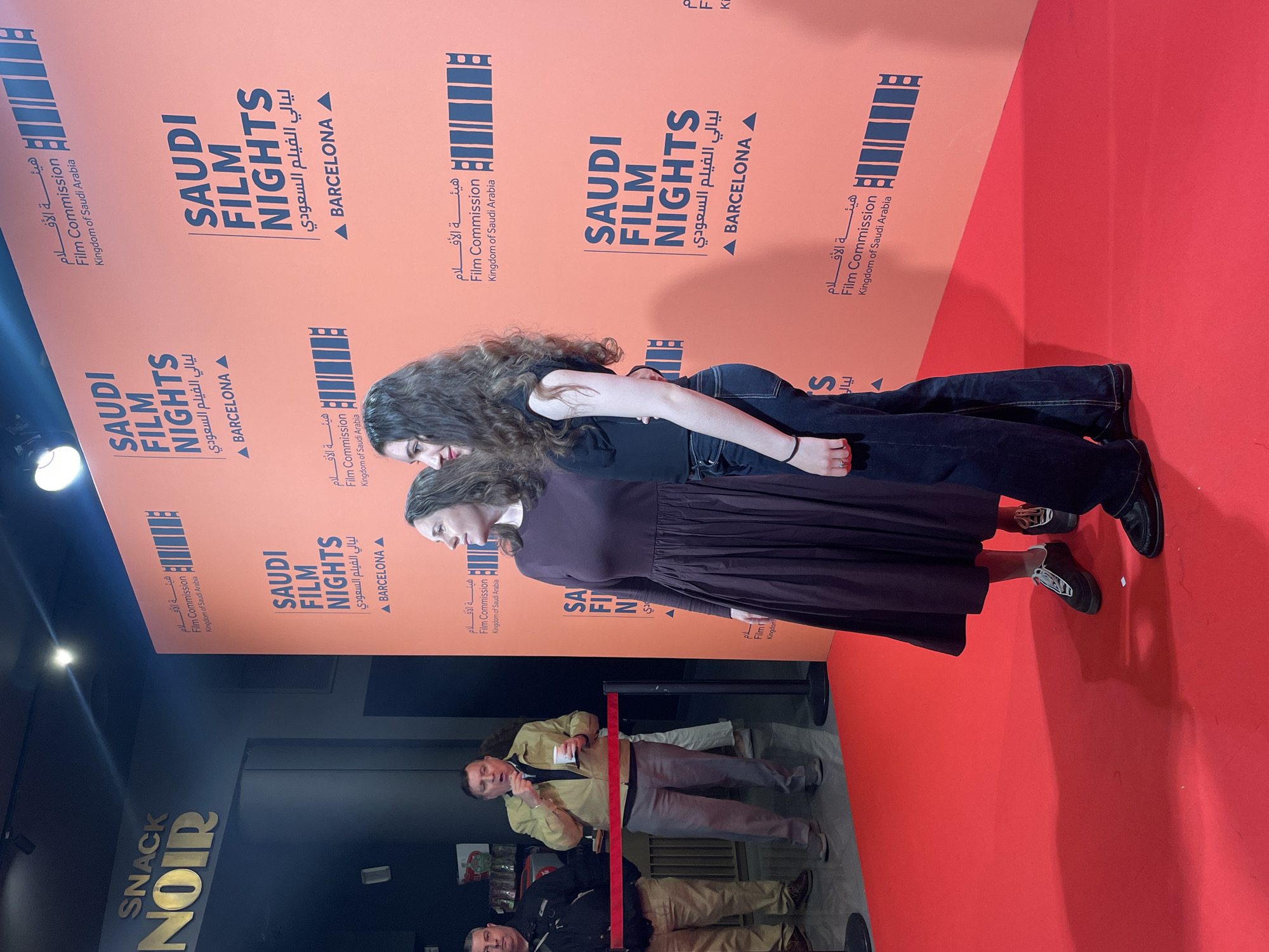Two women are standing in front of a red backdrop at what appears to be the "Saudi Film Nights" event in Barcelona, Spain. The backdrop features the event's logo and branding. One woman is wearing a brown dress, and the other is wearing a black top and jeans. They are standing close to each other, seemingly posing for a photograph. Behind them, a man in uniform is standing next to a red rope. "Snack Noir" is visible.