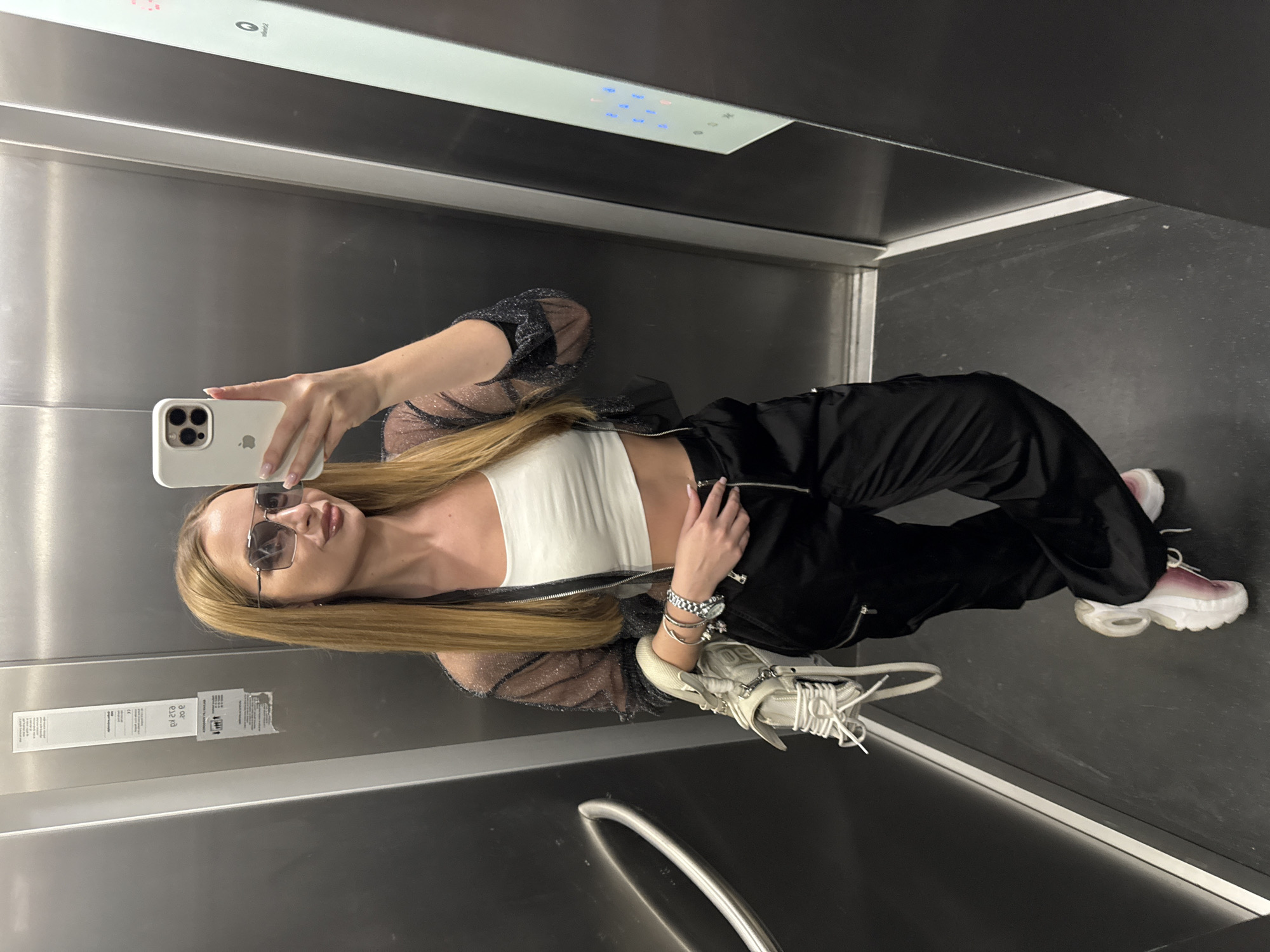 A young woman is captured in a selfie taken within an elevator, reflecting her image in the polished metallic wall. The image is oriented with the woman standing upright. She has long, straight blonde hair and is wearing rectangular-framed sunglasses. Her right hand holds a white iPhone, encased in a white cover, as she takes the photograph.
She is dressed in a white crop top, layered with a sheer, sparkly black long-sleeved top or jacket. Her lower body is clad in black pants, which feature a visible zipper detail. On her left wrist, she wears a silver watch and several silver bracelets. She completes her outfit with chunky white sneakers that have pink accents, and a white handbag with prominent laces is held or draped over her left arm.
The elevator interior features brushed metallic walls and a dark floor. Above her head, an elevator control panel is visible, displaying illuminated buttons in blue and a reddish/pink hue, along with what appears to be a "P" (possibly for park or floor indicator) symbol. On the left side of the frame, a white paper sign with unreadable black text is affixed to the wall, appearing mirrored and upside down due to the reflection. A silver handrail is partially visible at the bottom of the image. The scene takes place indoors, likely in Budva, Montenegro.
