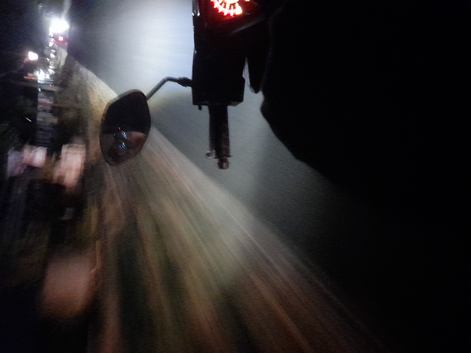 The image captures a first-person perspective from a moving vehicle, likely a motorcycle, at night on a street in Jetaksari, Indonesia. The foreground is dominated by the dark silhouette of the rider's hand and the handle of the vehicle, with a glowing red instrument panel visible on the right. The mirror of the vehicle reflects a blurred image of the rider and the road ahead. The road surface is illuminated by the vehicle's headlights, creating streaks of light that emphasize motion. To the left, the environment appears to be a roadside with some vegetation and indistinct structures. In the distance, streetlights and the headlights of other vehicles suggest an urban or semi-urban setting. The overall impression is one of night travel through a potentially dimly lit street.