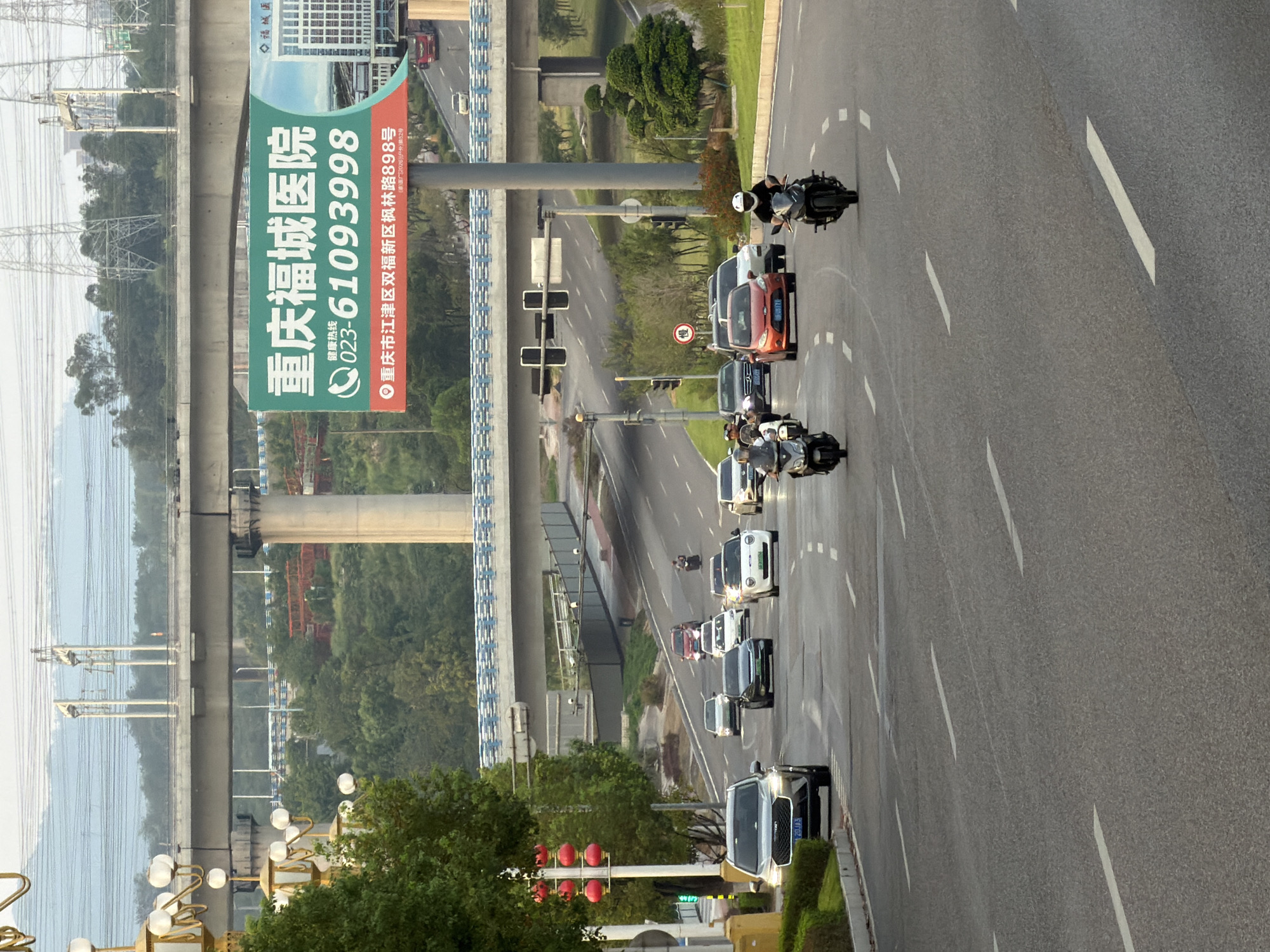 The image captures a street scene in Jiangjin District, Chongqing, China, characterized by elevated highways and urban infrastructure. The foreground features a multi-lane asphalt road with several vehicles, including cars and motorcycles, in motion. A gray SUV is prominent on the left, its license plate partially visible. Several motorcycles are dispersed throughout the lanes, with riders wearing helmets. In the middle ground, an orange car is visible, along with other cars and motorcycles. The road is lined with green vegetation and streetlights.
Dominating the background is a large billboard advertising "Chongqing Fucheng Hospital" with a phone number and address. Above the billboard, an elevated highway structure with supporting pillars and overhead wires is visible, suggesting the presence of a railway or power lines. Trees and buildings are scattered in the distance, forming a backdrop to the urban landscape. The sky is overcast, with the overall lighting indicating it is daytime. A distinct streetlight with red lanterns stands to the left of the road. The scene depicts a typical urban commute in a developed area of China.