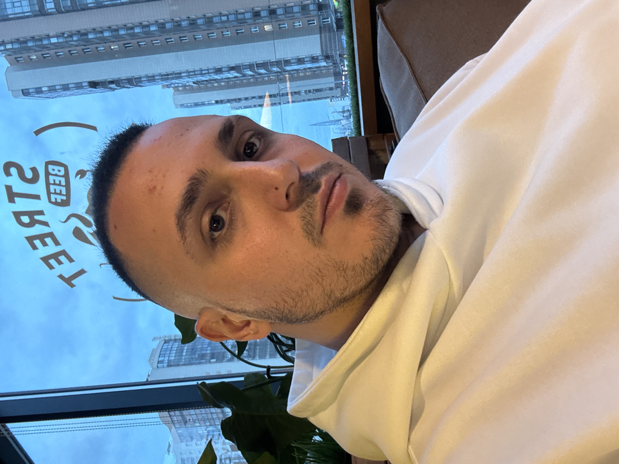 A close-up, slightly angled selfie of a man with short dark hair and a beard is captured indoors. He is wearing a white hoodie and is seated in a brown upholstered chair. Behind him, through a large window, a cityscape is visible. A building with multiple windows is reflected in the window. The reflection also shows a cloudy sky. A decorative plant is present in the foreground near the window. Part of a sign with the word "BEER" is visible, also reflected in the window. The city is Saint Petersburg, Russia.