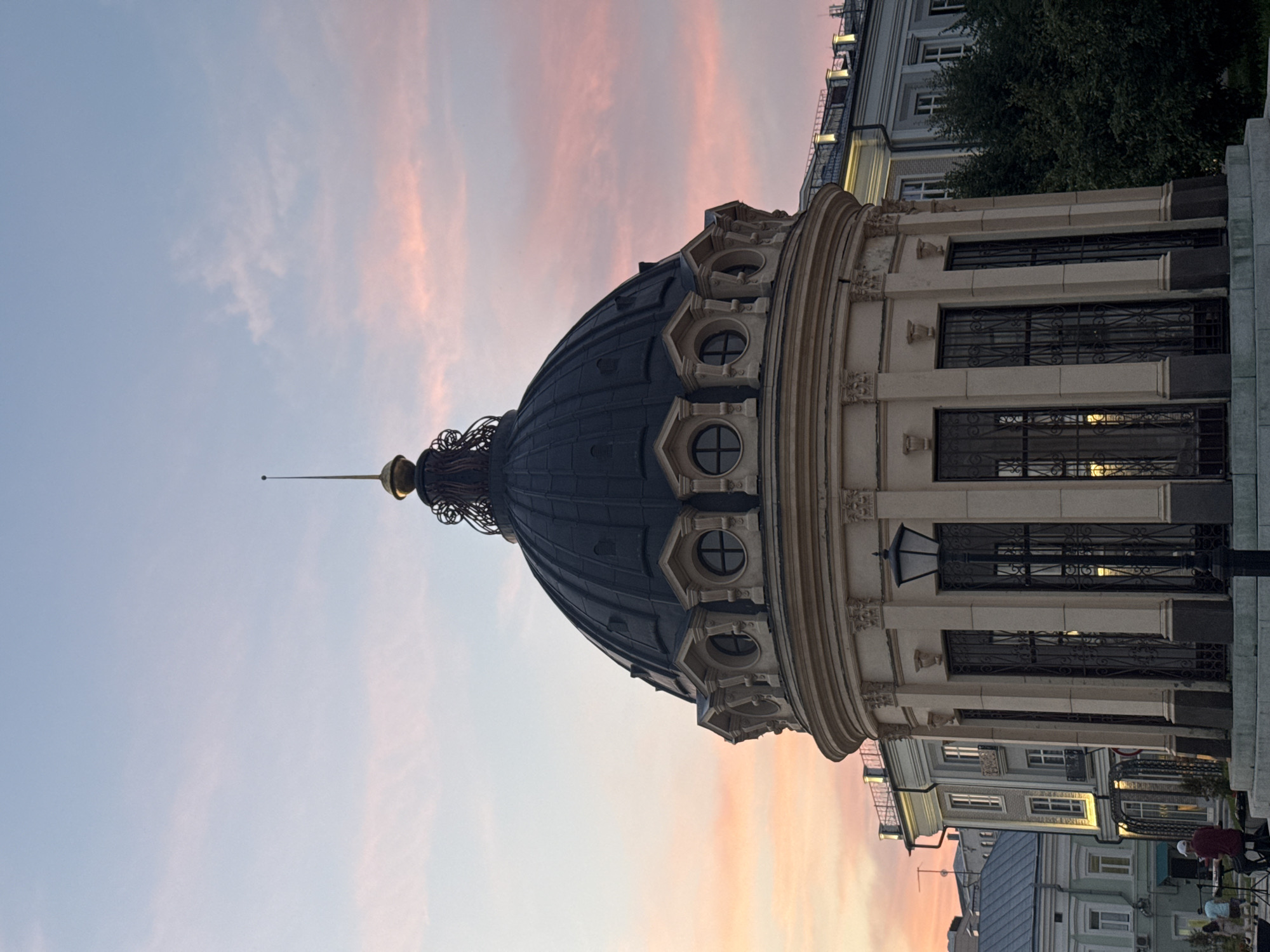 This is a vertical, eye-level, medium shot photograph of the Kazan Family Center, also known as the "Chalice," situated in Kazan, Russia. The architectural structure is a prominent domed building with a beige-colored facade adorned with columns and decorative elements. Above the facade is a dark, rounded dome topped with a golden spire. The sky in the background displays hues of pink and pale blue, suggesting it is either sunrise or sunset. In the lower left of the frame, two individuals are visible, one appearing to be playing a keyboard and the other standing nearby. The scene is set in an urban environment with buildings and trees in the background.