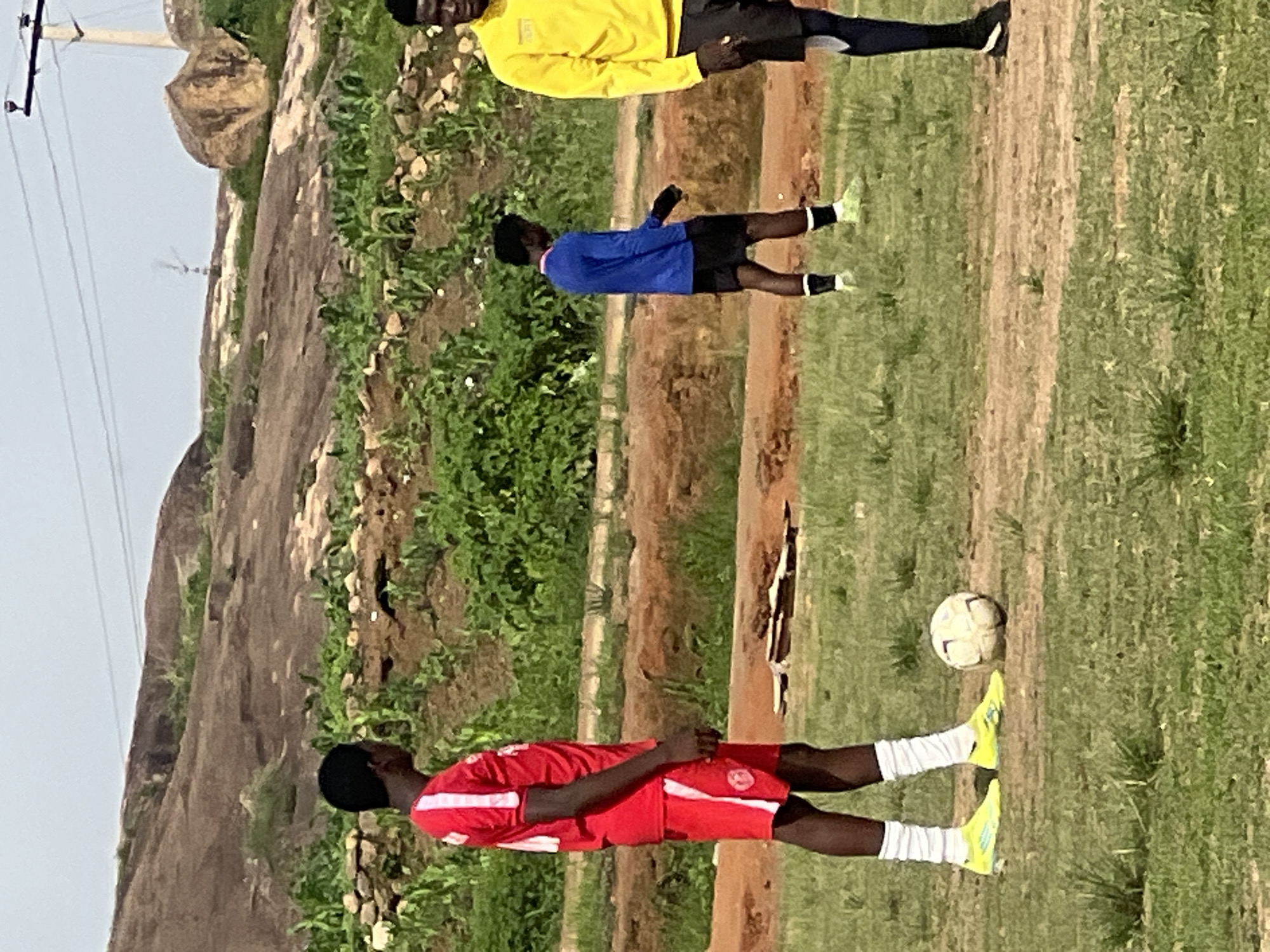 In a scene on the outskirts of Jos, Nigeria, three young men are engaged in a soccer practice session. The foreground is dominated by a grassy field with patches of bare earth. To the left, a man in a red soccer jersey and shorts stands with a soccer ball at his feet, his bright yellow cleats standing out against the green grass. His white socks are pulled up high. In the middle ground, a man in a blue long-sleeved shirt and black shorts walks away from the camera, also wearing light-colored cleats and white socks. To the right, another man, partially visible, wears a bright yellow long-sleeved shirt and dark shorts.
The background reveals a hilly landscape with large rock formations and sparse vegetation, suggesting a semi-arid or rural environment. A power pole with wires is visible in the upper right corner, indicating the presence of infrastructure. The sky is overcast, casting a diffused light on the scene. The overall impression is of an informal outdoor sports activity in a somewhat rugged natural setting.
