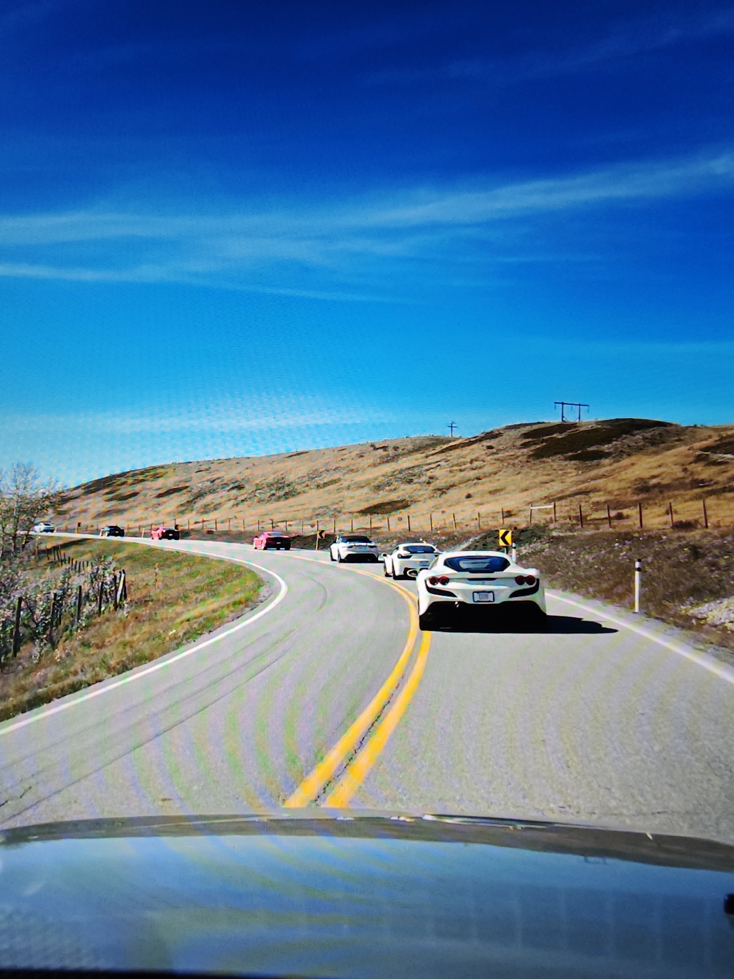 The image depicts a convoy of sports cars driving on a winding paved road in a rural, hilly area with sparse vegetation under a bright blue sky. The lead car in the foreground is a white sports car, followed by several other sports cars, including two brightly colored pink ones. The road has double yellow lines indicating a no-passing zone. In the background, there is a dry, grassy hill with some fencing. The scene appears to be a scenic drive, possibly in an area outside of Calgary, Canada, known for its natural landscapes. The overall impression is one of leisure and automotive enjoyment in a picturesque natural setting.