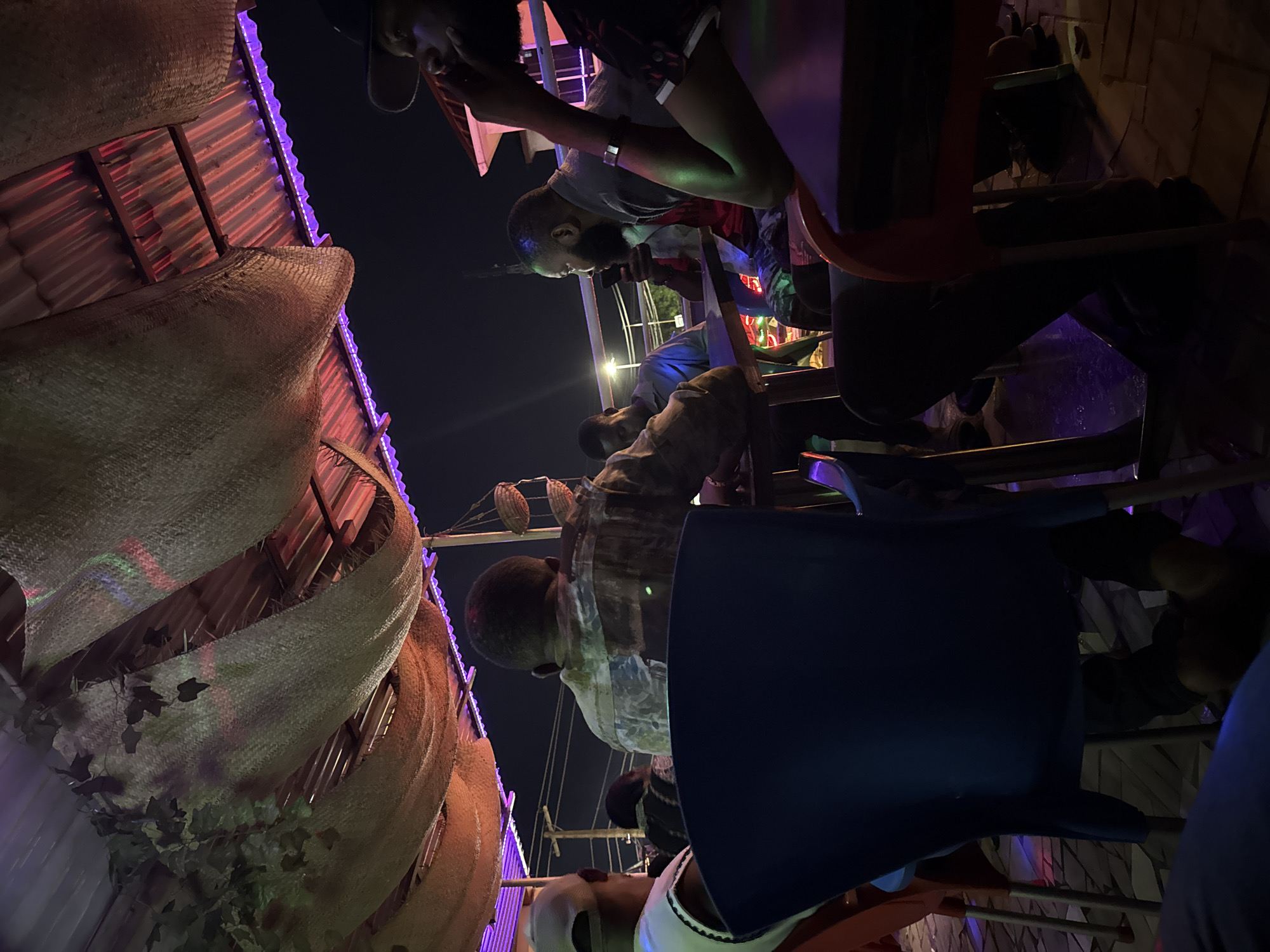 At night, a low-angle photograph, rotated counter-clockwise, shows a gathering of people beneath a structure with thatched decorations and purple LED lighting. Seven individuals are discernible, seated on an assortment of blue and orange plastic chairs. One man in a grey shirt looks down at his phone, while another in a camouflage shirt sits nearby. A man in a white cap is visible in the foreground. The setting appears to be a casual outdoor venue, possibly a bar or restaurant, located in Ado, Nigeria.