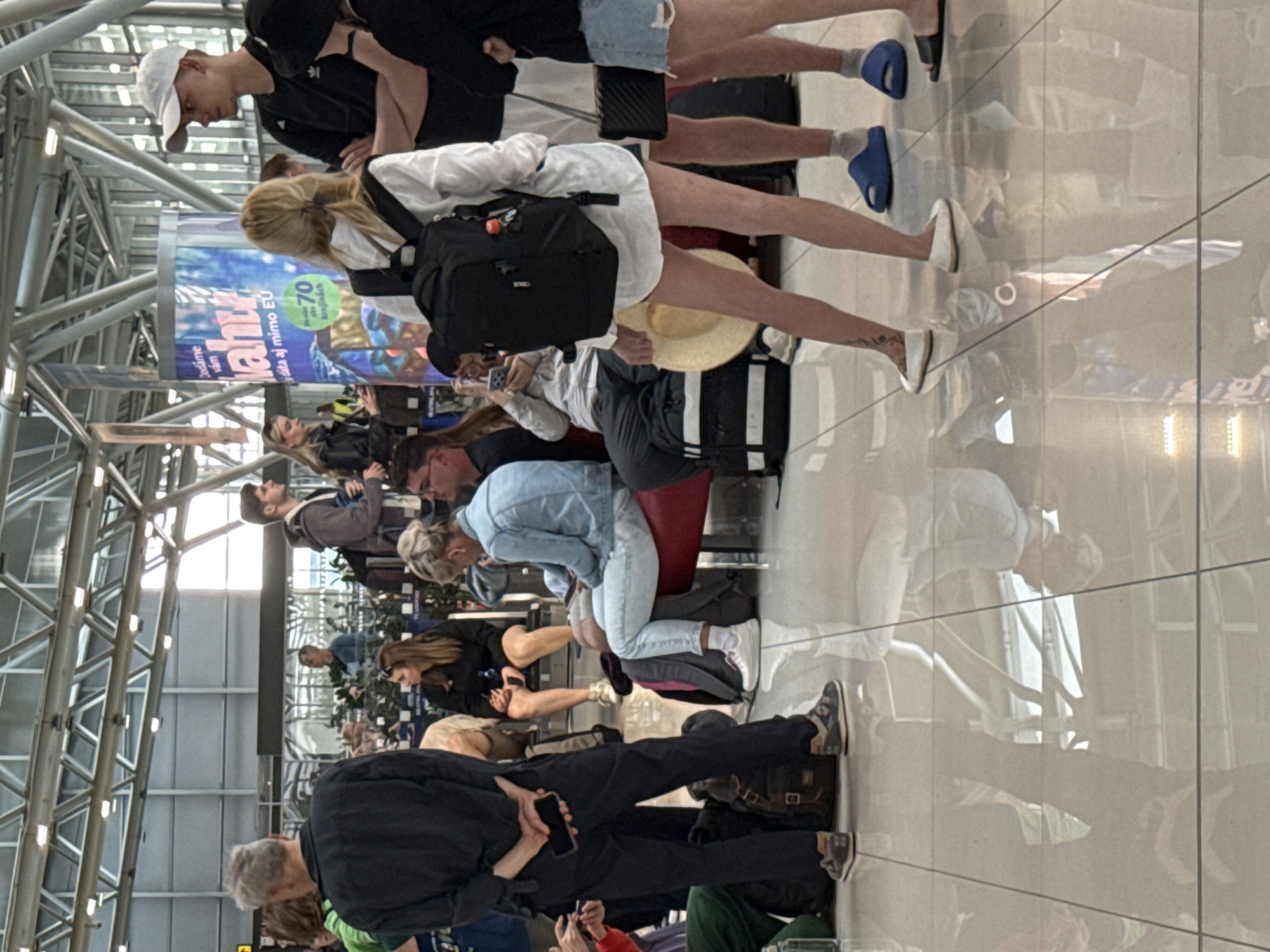 The image depicts a group of people inside an airport terminal in Bratislava, Slovakia. The environment is characterized by polished tile flooring that reflects the overhead structure and the individuals present. People are seen standing and sitting, many appearing to be waiting, with luggage visible around them.
In the foreground, a person from the waist down is wearing dark pants and a black jacket with their hands clasped behind their back, holding a smartphone. A backpack is on the floor to their left. To their right, a woman in a white shirt and black backpack is standing, holding a straw hat. Further to the right, two people are partially visible, one wearing a denim skirt and the other blue slides.
In the mid-ground, a woman in a blue denim jacket and light-colored pants is sitting on a red bench, looking at her phone. To her left, a man in a light blue shirt is sitting beside her. Behind them, other individuals are seated on benches and standing, some with backpacks.
A large cylindrical advertisement featuring a blue and white design with text in Slovak is positioned behind the people in the mid-ground. The overall scene suggests a typical airport waiting area.