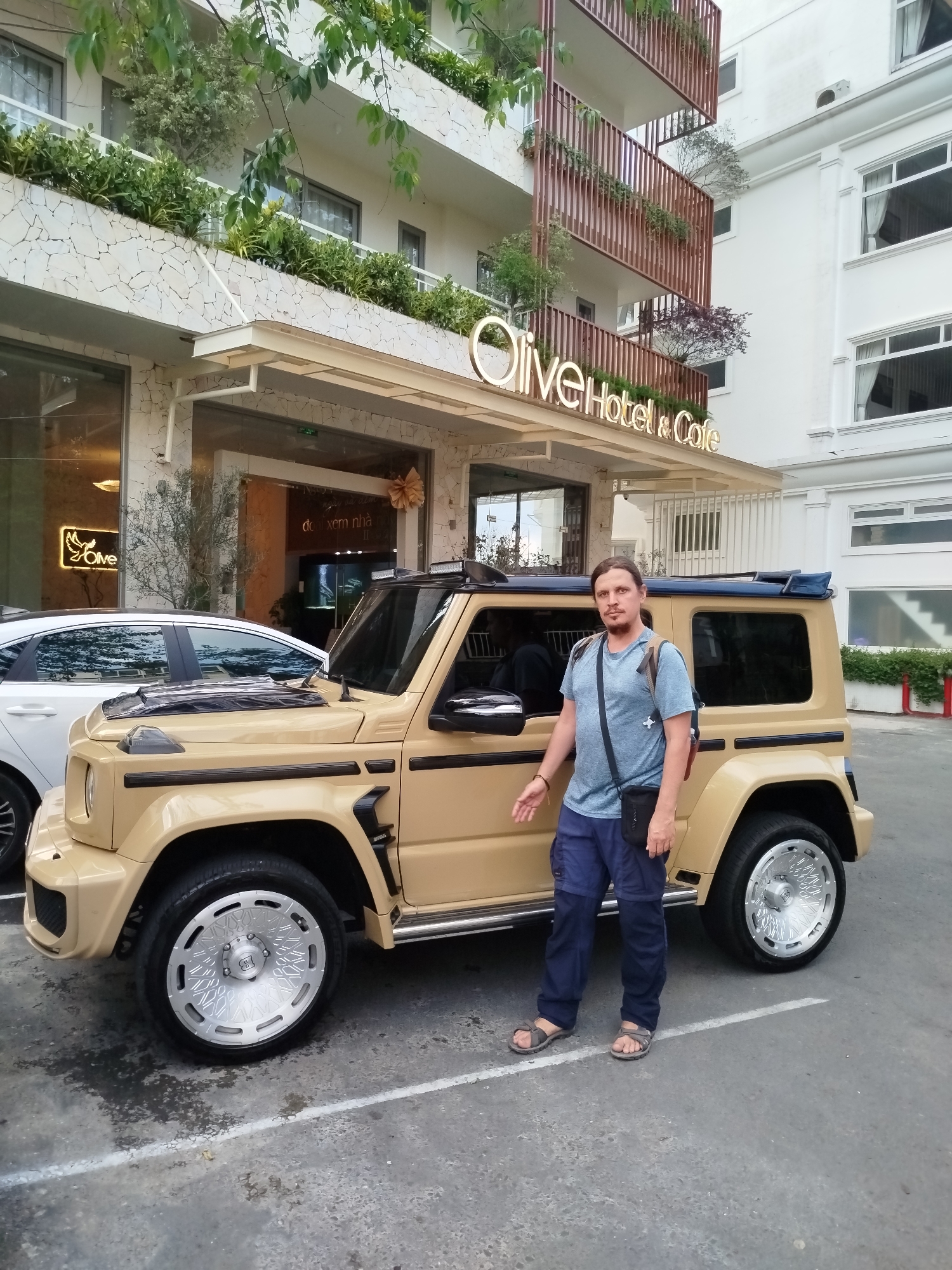 The image captures a man standing next to a modified, tan-colored Suzuki Jimny vehicle in front of the "Olive Hotel & Cafe." The hotel is a multi-story building with a stone façade and decorative greenery. The man is wearing a blue t-shirt, dark blue pants, and sandals. He has shoulder-length brown hair and a small bag slung across his shoulder. The Suzuki Jimny is customized with aftermarket wheels and body modifications. A white sedan is partially visible behind the Jimny. The location is likely Da Lat, Vietnam, indicated by the building style and local businesses in the background. The scene suggests an outdoor parking area or driveway in front of the hotel.